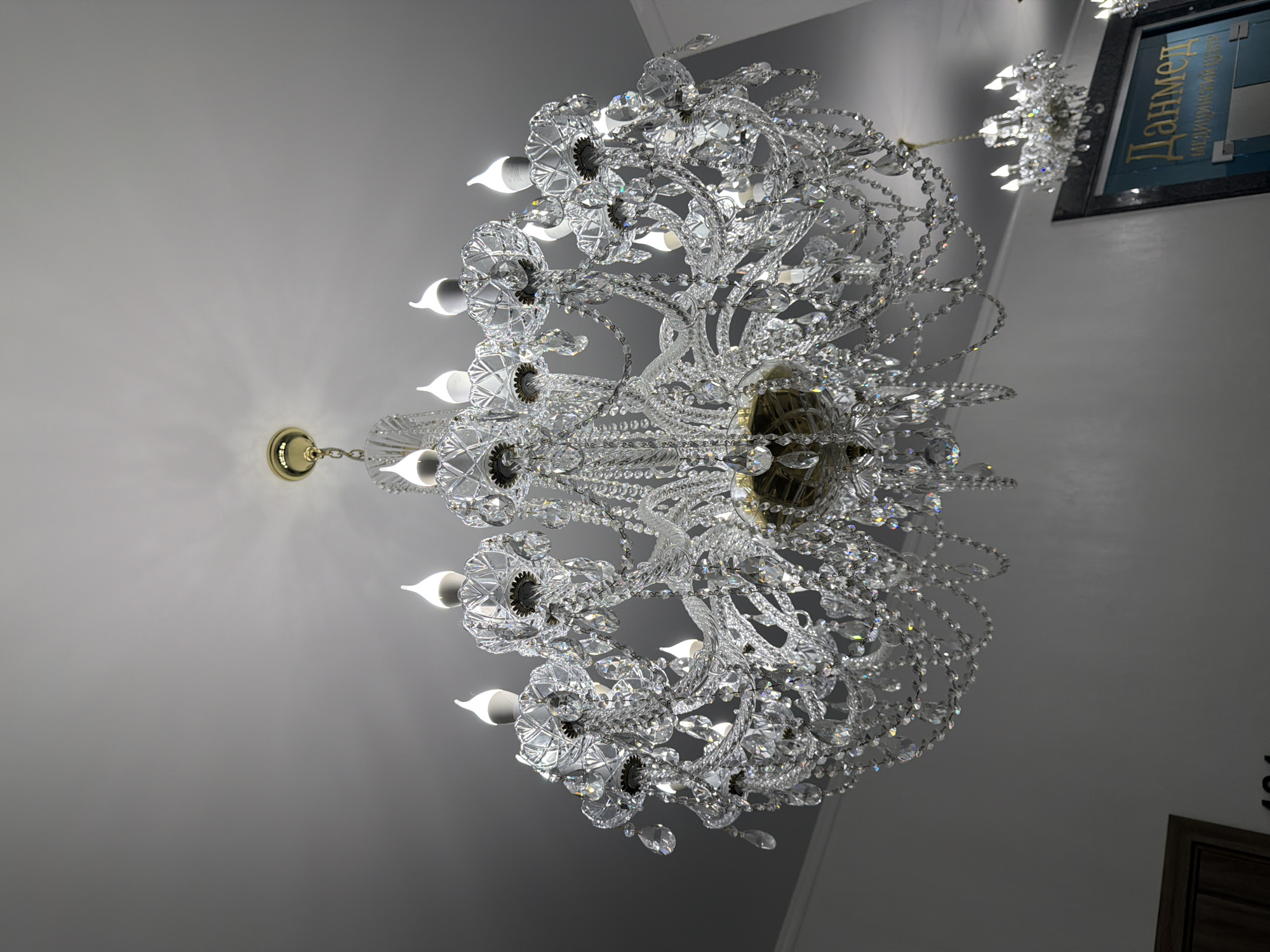 The media file displays a lavish crystal chandelier hanging from a white ceiling. The chandelier is ornate, with multiple tiers of sparkling crystals and delicate metalwork, resembling branches adorned with leaves and droplets. Numerous light bulbs, shaped like candles, are incorporated into the design, casting a warm glow.
In the background, a framed sign is visible on the wall, featuring text in Russian. The text appears to be "Данмед" in large, stylized letters, with smaller text below it. The overall impression is one of opulence and elegance, likely an interior setting such as a hotel, restaurant, or a well-decorated home. Given the location context of Sochi, Russia, it could be a high-end establishment. There are no people visible in the image, and the focus is entirely on the decorative lighting fixture and the accompanying sign. The lighting suggests it might be evening or an indoor setting with controlled lighting.