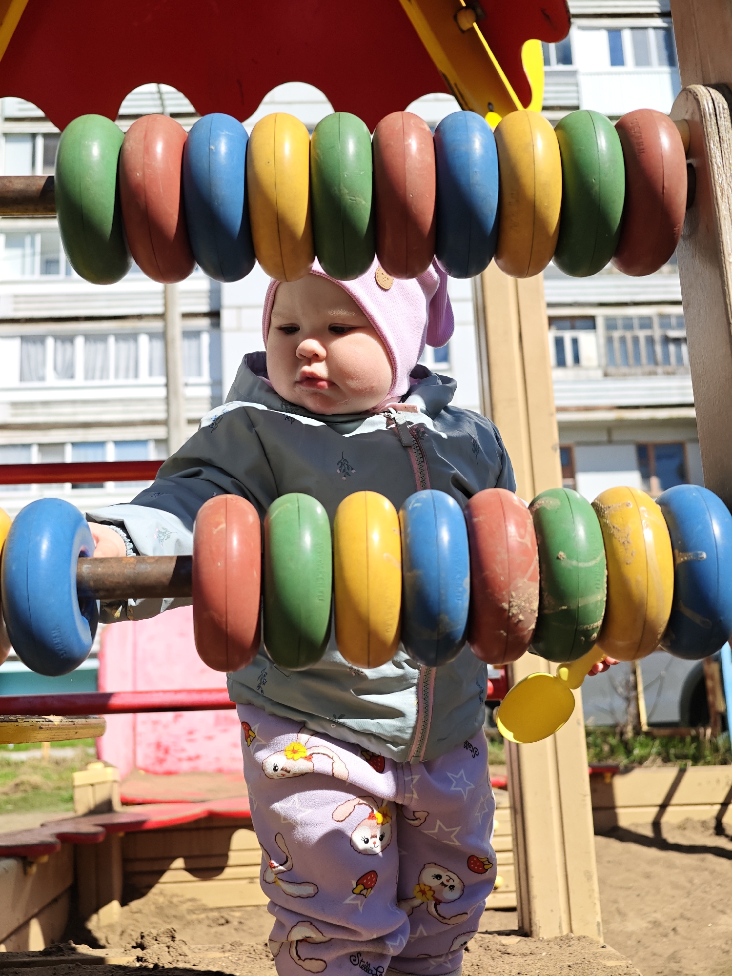 A young child stands at a playground apparatus consisting of two horizontal bars strung with large, colorful beads in the city of Chaikovsky, Russia. The child is wearing a grey jacket with a hood, and patterned lilac pants with cartoon bunny faces and strawberry graphics. The child's face is turned to their left, and their right hand is touching the beads on the lower bar, with a yellow plastic scoop in their grip. The beads are arranged in a repeating color sequence of green, brown, yellow, and blue. The playground equipment is set against a background of a large apartment building with white window frames. The ground around the playground is covered in sand.