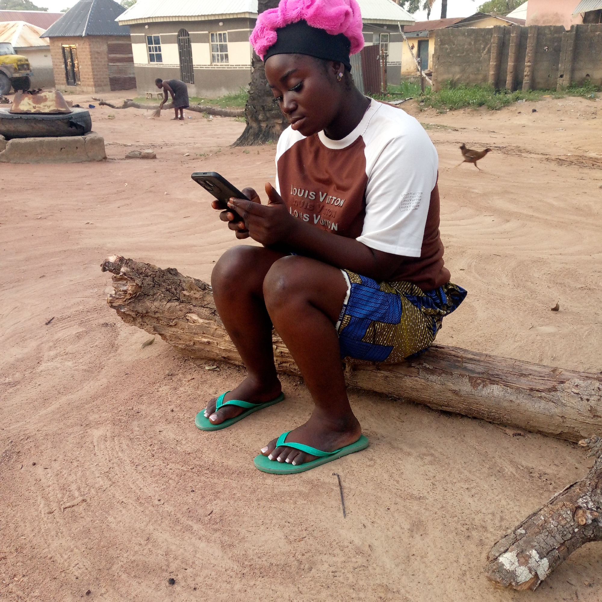 A young woman with dark skin and a pink head covering sits on a log outdoors, using a smartphone. She wears a brown and white shirt, a colorful patterned skirt, and green flip-flops. The ground is sandy and there is a nail in the ground near her feet. In the background are simple buildings, a person sweeping, and a chicken. The location is Mararaba, Nigeria.
