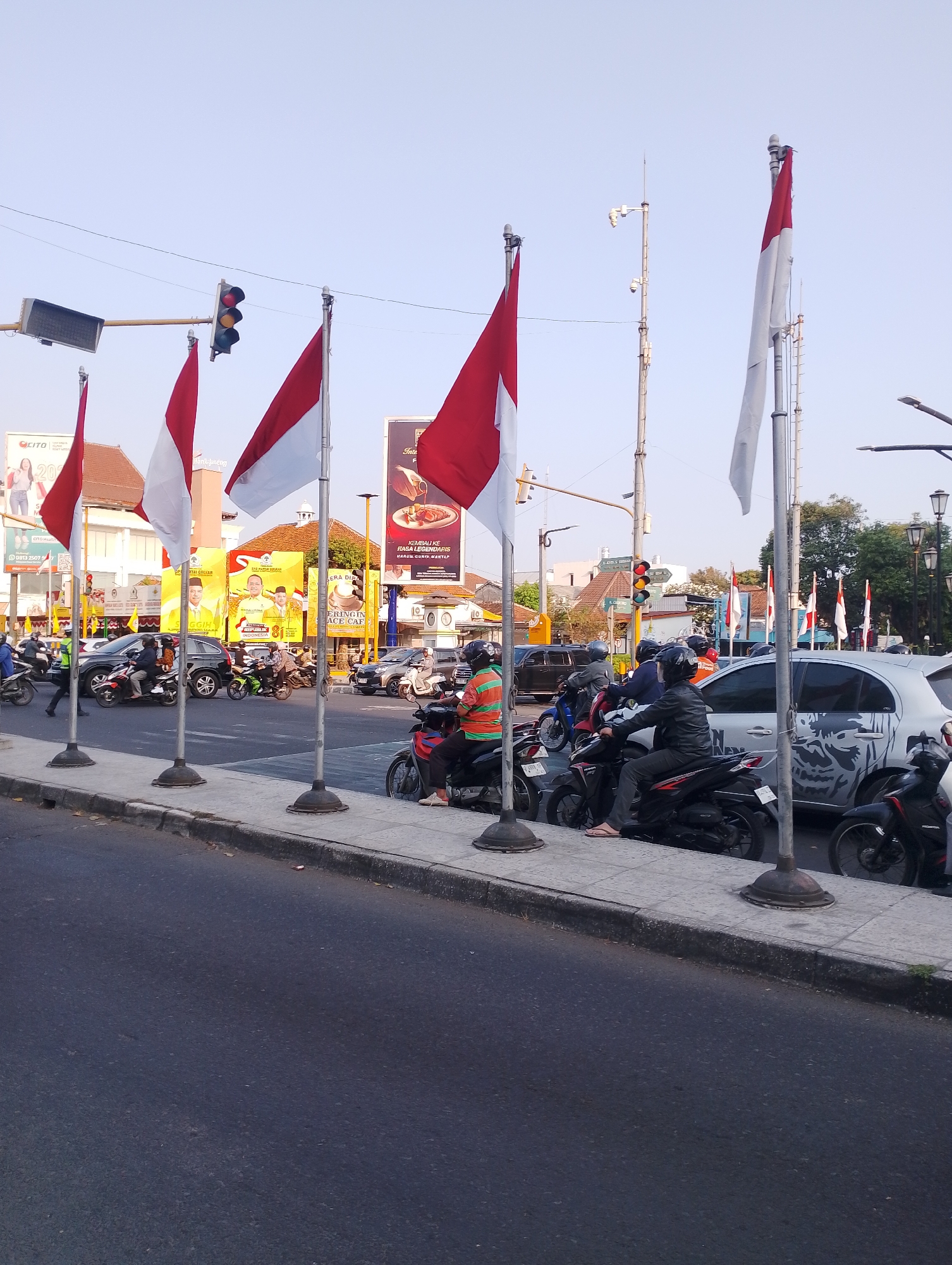 This image captures a street scene in Yogyakarta, Indonesia, likely near a traffic intersection given the presence of traffic lights and a busy road. The foreground shows a sidewalk lined with several Indonesian flags on poles. Beyond the sidewalk, traffic is visible, including motorcycles and cars. People are riding on the motorcycles, some wearing helmets. In the background, buildings and billboards are visible. The sky is clear and the scene appears to be during daylight hours.