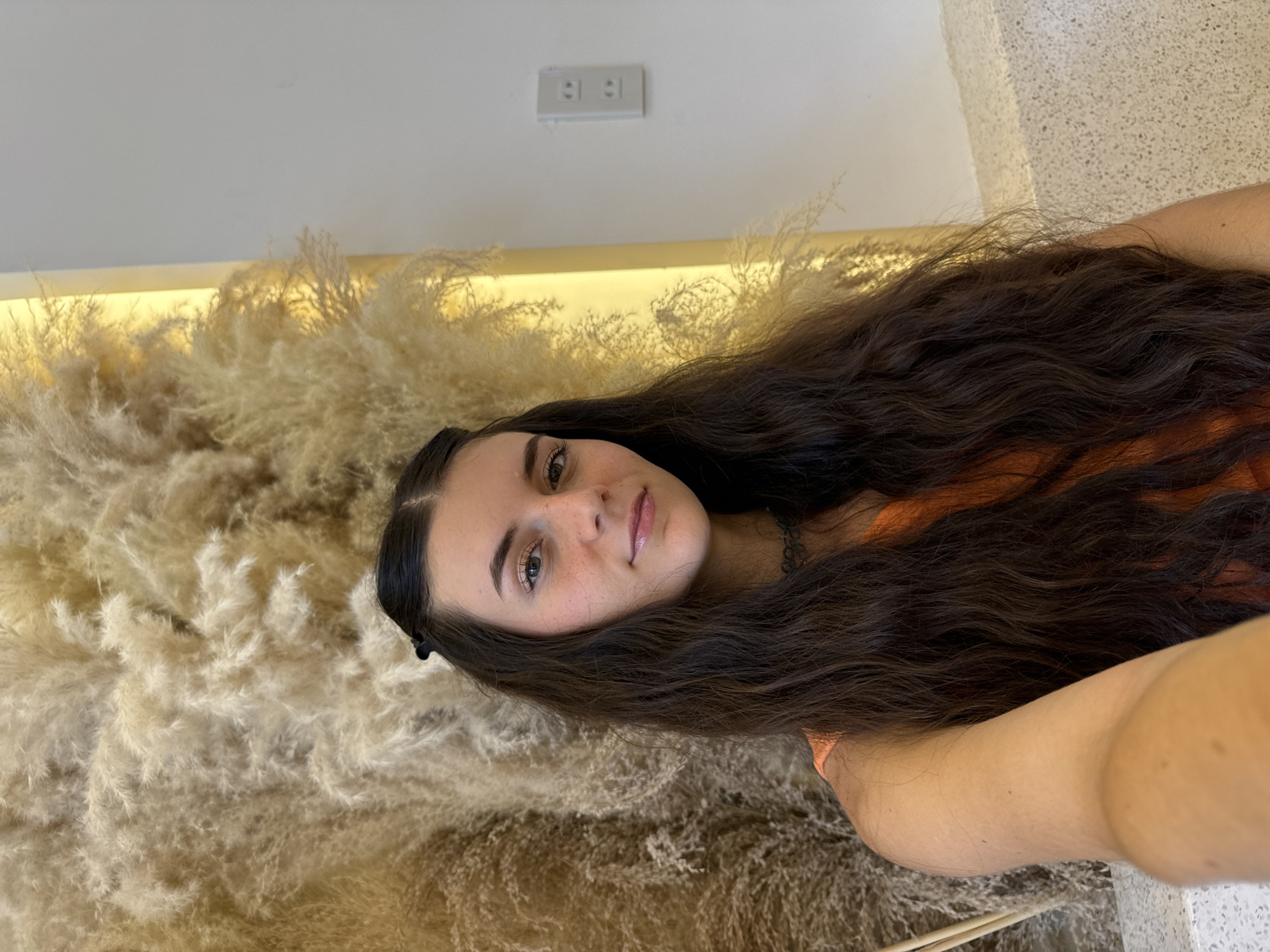 The image presents a young woman in a close-up portrait, captured indoors in Phu Thuy Ward, Vietnam. The photograph is taken with the camera rotated, positioning her head towards the top left of the frame. She has long, dark brown, wavy hair that cascades downwards and to the right. Her fair skin is adorned with visible freckles across her nose and cheeks, and she has light-colored eyes. She is wearing a light orange top and a dark choker necklace, exhibiting a calm, slightly smiling expression as she looks directly at the camera.
The background on the left and bottom is largely dominated by a voluminous arrangement of light brown or beige pampas grass, creating a soft, feathery texture. Above this natural decor, a warm yellow strip of light illuminates a smooth, light-colored wall. A white electrical outlet with two horizontal sockets is mounted on this wall in the upper center of the image. A subtly textured, light-colored surface is visible on the far right, possibly another wall or furnishing. The setting suggests a modern and stylish interior environment, potentially a cafe or a contemporary living space. No visible text is present in the image.