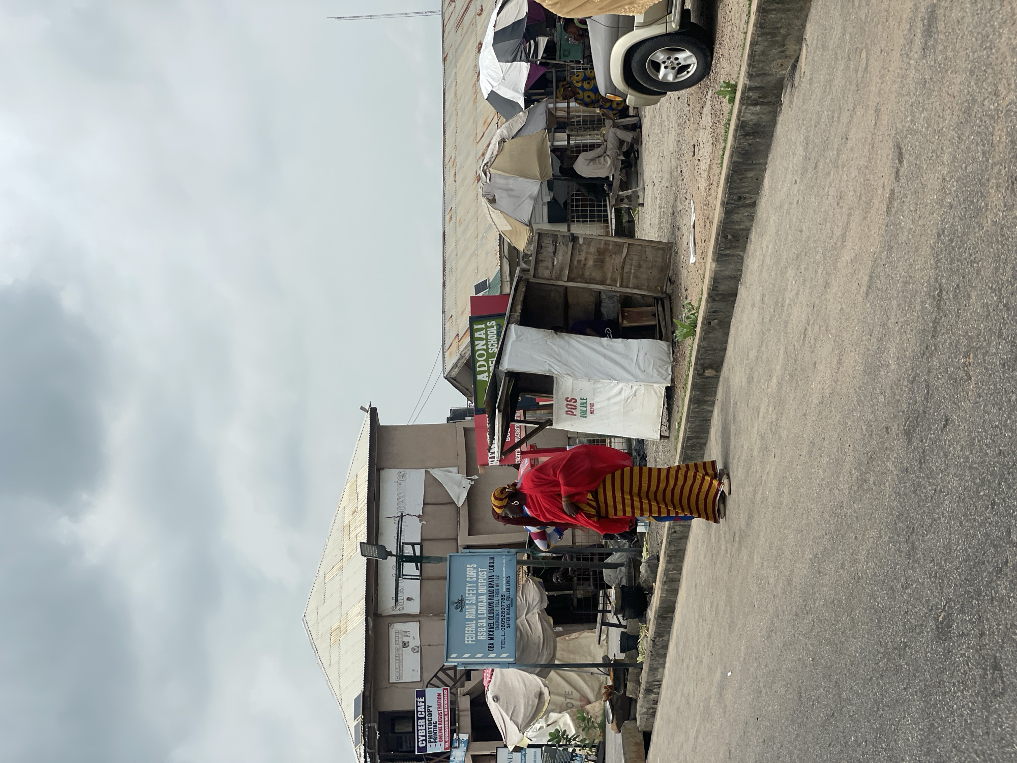 The image depicts a street scene in Lokoja, Nigeria, under an overcast sky. In the foreground, a paved road runs diagonally from the bottom left to the top right. Along the right edge of the road, a concrete curb separates the pavement from a dirt area. On the dirt area, a beige-colored SUV is partially visible on the right, with its front wheel and fender on display.
To the left of the SUV, a collection of market stalls are set up. One stall features a wooden structure with an open front, covered by a white tarp. Next to it, a white and black umbrella shades another stall. Further to the left, several other stalls are visible, some covered by awnings or tarps.
Prominently displayed in the middle ground is a blue sign for the "FEDERAL ROAD SAFETY CORPS RS8.3A LOKOJA OUTPOST OBA MICHAEL GLOBAYO ROAD KPATA, LOKOJA". Below this, a red and yellow striped dress and a red head wrap are worn by a person facing away from the camera and towards the stalls, their hands resting on their waist. Another person is partially visible behind them.
In the background, a building with a corrugated metal roof is situated behind the market stalls. Signs are attached to the building, including one that reads "CYBER CAFE PHOTOCOPY" and another with "COSMETICS SHOP". To the right of this building, another structure with a similar corrugated roof is visible, and a sign with green lettering reads "ADONAI SCHOOLS". The overall environment suggests a busy, informal market area in Lokoja.