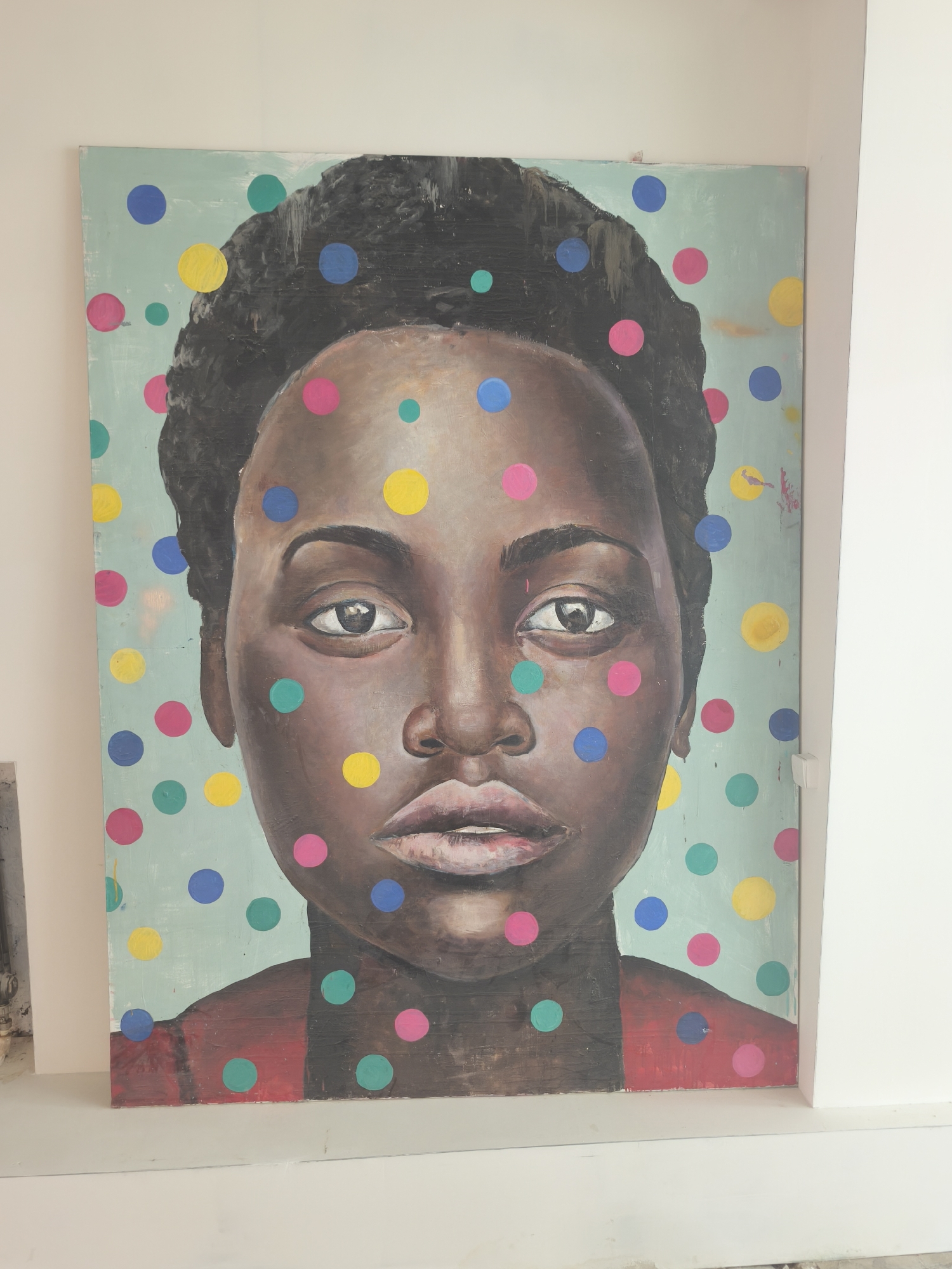 This is a painting of a young Black person's face, rendered in a realistic style against a pale teal background dotted with colorful circles. The subject has dark, curly hair, prominent eyes with reflective pupils, and full lips painted in a realistic shade. The dots, in various shades of yellow, pink, blue, and teal, are scattered across the canvas, some overlaying the subject's face and hair, adding a whimsical and abstract element to the portrait. The subject is depicted from the chest up, with a dark neckline suggesting clothing. The overall mood is somewhat somber due to the subject's direct gaze and expression, contrasted with the playful nature of the polka dots. The painting appears to be on a canvas or wood panel, with visible brushstrokes and some signs of age or wear on the edges of the work. The setting appears to be indoors, possibly an art studio or exhibition space, with plain white walls and a ledge beneath the artwork. There is no visible text in the image.