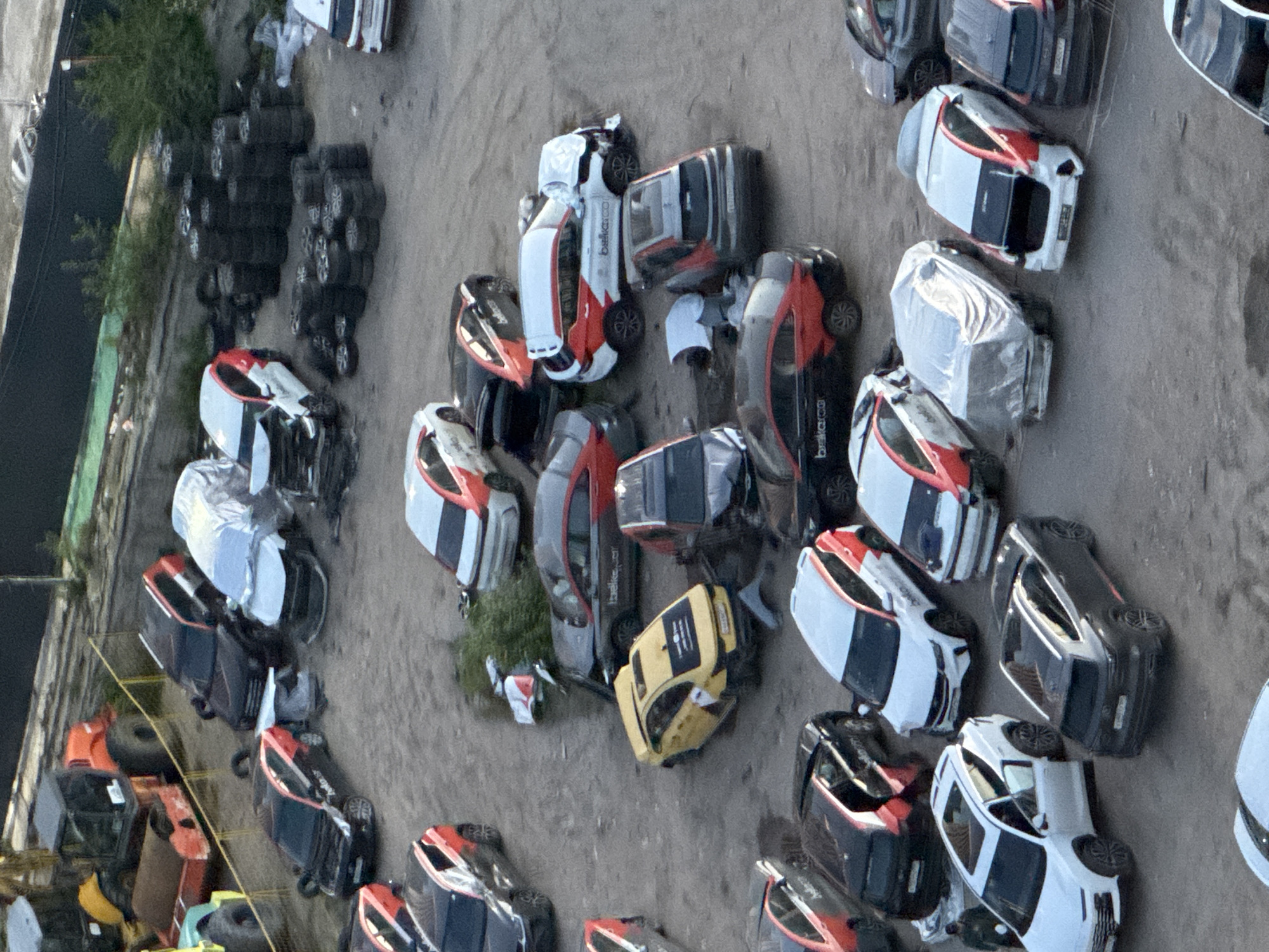 An aerial view shows a large outdoor impound lot filled with numerous damaged and derelict vehicles. The lot appears to be located in Bobrovo, Russia, indicated by markings on some of the cars. The majority of the vehicles are white and red, suggesting they may be part of a fleet. Several cars are visibly damaged, with crumpled bodies and missing parts, indicating they have been involved in accidents. Some vehicles are covered with tarps. In the background, construction equipment, including a bulldozer and a roller, are present. A large stack of tires is also visible in the middle of the lot. There are no people clearly visible in the scene. The ground is unpaved and dusty, typical of an outdoor storage or impound area.