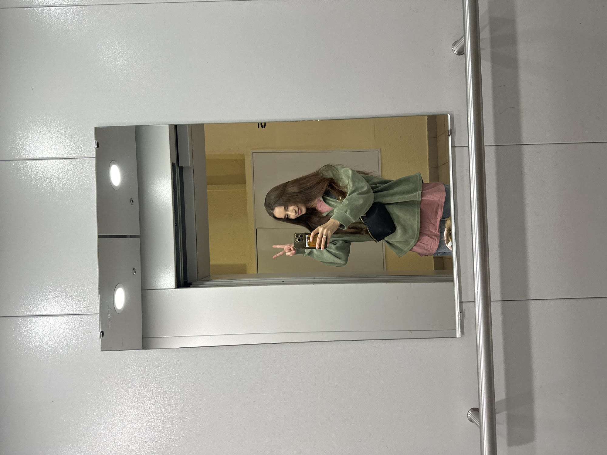 A young woman takes a mirror selfie in a brightly lit elevator. She's smiling and holding up two fingers in a peace sign. She is wearing a pink top, a light green jacket, and a black bag. The elevator is modern with a light gray interior and features a vertical metal handrail. A sign that says '10' is visible at the top of the mirror. The location is likely in Yekaterinburg, Russia.