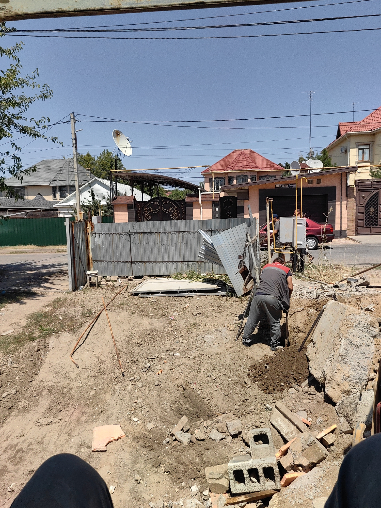 In Shymkent, Kazakhstan, on a clear day, construction work is underway in a dirt lot adjacent to a street. A man in gray work pants and a grey shirt with red accents is digging a trench with a shovel. Piles of dirt and construction debris, including concrete blocks and broken bricks, surround the excavation site. A corrugated metal fence and gate are visible behind the construction area, with a detached section of corrugated metal leaning against it. In the background, houses with red-tiled roofs and a car are present, along with utility poles and satellite dishes. A green metal fence runs along the street. The overall scene depicts an active construction or renovation project in a residential urban environment.