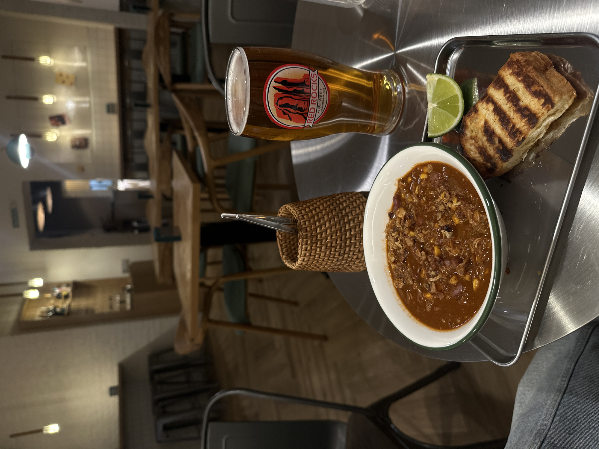 The image depicts a meal at what appears to be a restaurant. The focus is on a metal tray containing a bowl of chili, a lime wedge, and grilled bread. Beside the tray, there's a glass of beer with the "Red Rocks Brewery" logo and a woven container holding silverware. The setting appears to be a restaurant interior, with wooden tables and chairs visible in the blurred background. Based on the beer label (Red Rocks Brewery), the image might be taken in Krasnoyarsk, Russia where the brewery is located.