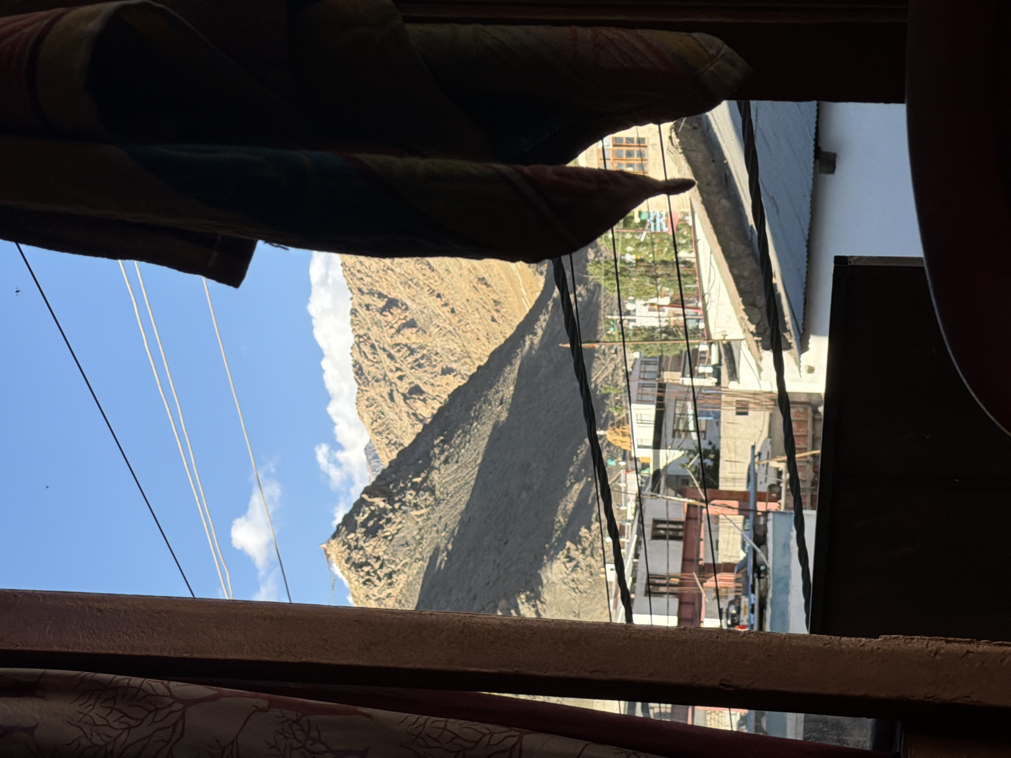The image presents a view through a window frame, with a curtain on the left and a draped fabric on the right partially obscuring the scene. The view looks out onto a town with buildings and a mountainous backdrop, characteristic of Leh, India. In the foreground, the window frame and the dark shape of furniture are visible. Beyond these, a town with multi-story buildings, some with traditional architecture featuring wooden window frames, is seen. Cars are parked on a street or courtyard within the town. Above the buildings, several power lines crisscross the sky. The background is dominated by rugged, sun-drenched mountains, with a clear blue sky dotted with a few white clouds. The overall lighting suggests daytime with strong sunlight casting shadows. The drapery on the right has visible striped patterns.