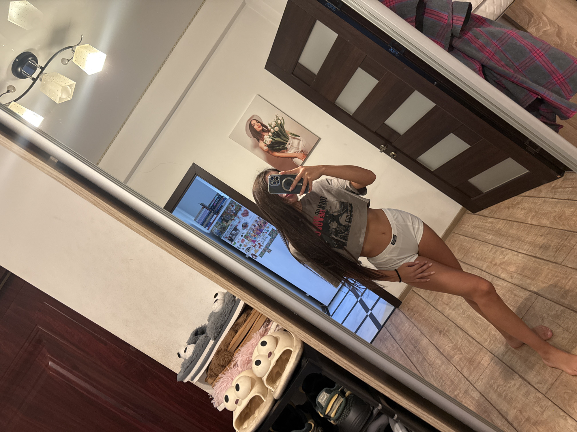 A young woman with long brown hair is taking a selfie in a mirrored closet door. She is wearing a grey crop top with a graphic print and white shorts. She is standing with her legs crossed and her right hand resting on her hip. In the reflection, we can see a painting of a woman holding flowers on the wall, a wooden door with glass panels, and a ceiling light fixture. Below the woman, in the closet, there are shelves with slippers and shoes. The floor is made of wood planks. The overall scene appears to be an indoor setting, possibly a bedroom. The image is taken with a phone, as indicated by the camera held in her hand. No text is clearly legible. The setting suggests a modern apartment in Chișinău, Moldova.