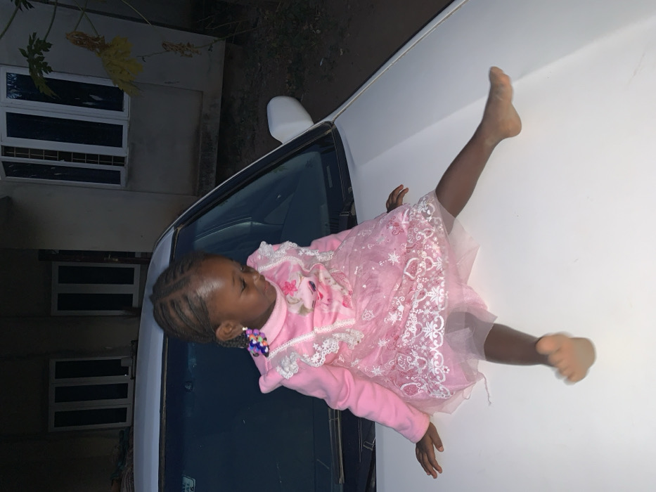 A young girl wearing a pink dress and jacket lies on the hood of a white car. Her bare feet extend towards the top right corner. The car's windshield and side mirror are visible. A building with gridded windows is in the background. Sparse plants and a hint of sky are also present, suggesting an outdoor setting. The image is slightly tilted.