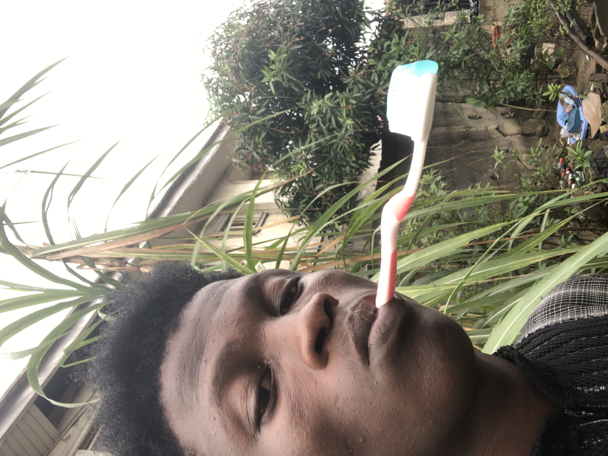 A close-up, waist-up, outdoor, eye-level, medium shot shows a young Black person in Ado, Nigeria. They are holding a white and pink toothbrush with blue bristles in their mouth, with the bristles against their lips. The person has dark, curly hair and is wearing a black and white patterned shirt with a black collar. Behind them, to the left, are tall green plants. To the right is a concrete wall with dense green foliage and trees. In the lower right corner, there is a blue plastic basin filled with debris, including bottles and trash. The sky is overcast and bright.