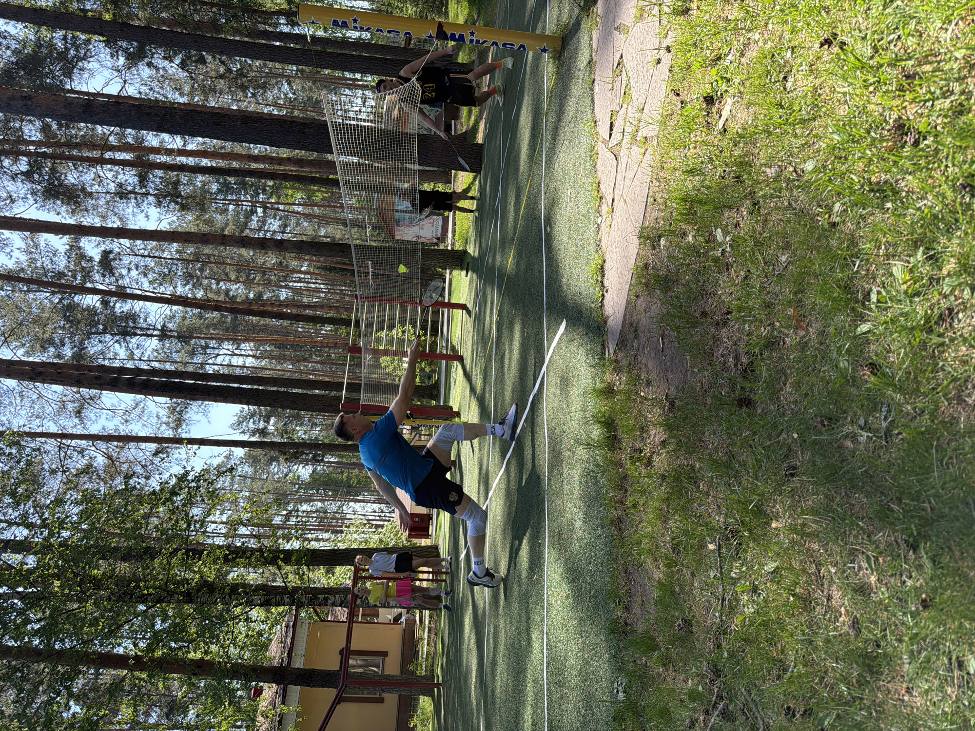 A sunny outdoor scene shows two men playing what appears to be speedminton on an artificial turf court surrounded by tall pine trees. The location is presumably in Chaykovsky, Russia, as per the prompt's instruction.
One man, in the foreground, is actively hitting the shuttlecock. He is wearing a blue shirt, black shorts, white knee supports, and grey sneakers. His arm is raised, indicating the moment of impact.
Across the net, another man wearing a black and yellow jersey (#24 visible) is mid-jump, likely preparing to hit the shuttlecock back.
The court is marked with white lines and has a net strung between yellow and blue Mikasa poles. There are other people, perhaps spectators, visible in the background. The ground adjacent to the court is a mixture of grass and what appears to be concrete paving stones. A small building with a yellow facade and dark roof is also visible in the background, partly obscured by trees.