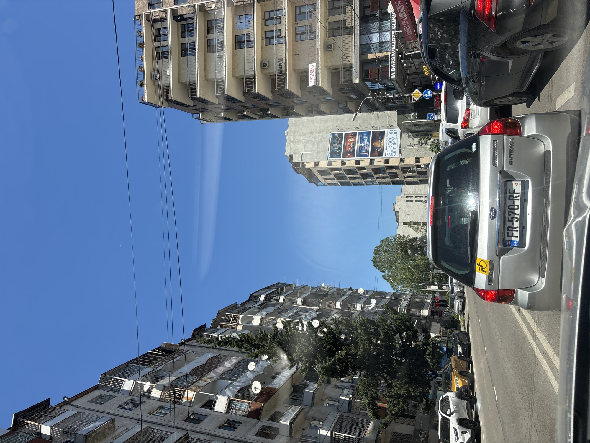 The image depicts a street scene in Tbilisi, Georgia, likely during daylight hours. The perspective is from inside a vehicle, facing forward in traffic. The road is paved, with visible lane markings. On either side of the street are multi-story apartment buildings with balconies and windows. Some of the buildings appear older and somewhat weathered, while others are more modern. Satellite dishes are visible on some of the older buildings.
Several cars are present in the frame, indicating a traffic jam or slow-moving traffic. The foreground shows the rear of a silver Subaru Outback with a yellow sticker on its rear window and a license plate that reads "GE FR-570-RF". To the right, the rear of a black SUV is visible. In the distance, more cars and buildings line the street.
A large billboard is prominently displayed on one of the buildings on the right side of the street, featuring images of interior designs and text in Georgian. Above the street, power lines are strung across, common in urban environments. The sky is clear and blue with some wispy clouds. Trees line parts of the street, adding greenery to the urban landscape. The overall impression is of a typical urban street in Tbilisi on a sunny day, characterized by traffic congestion and a mix of architectural styles.
