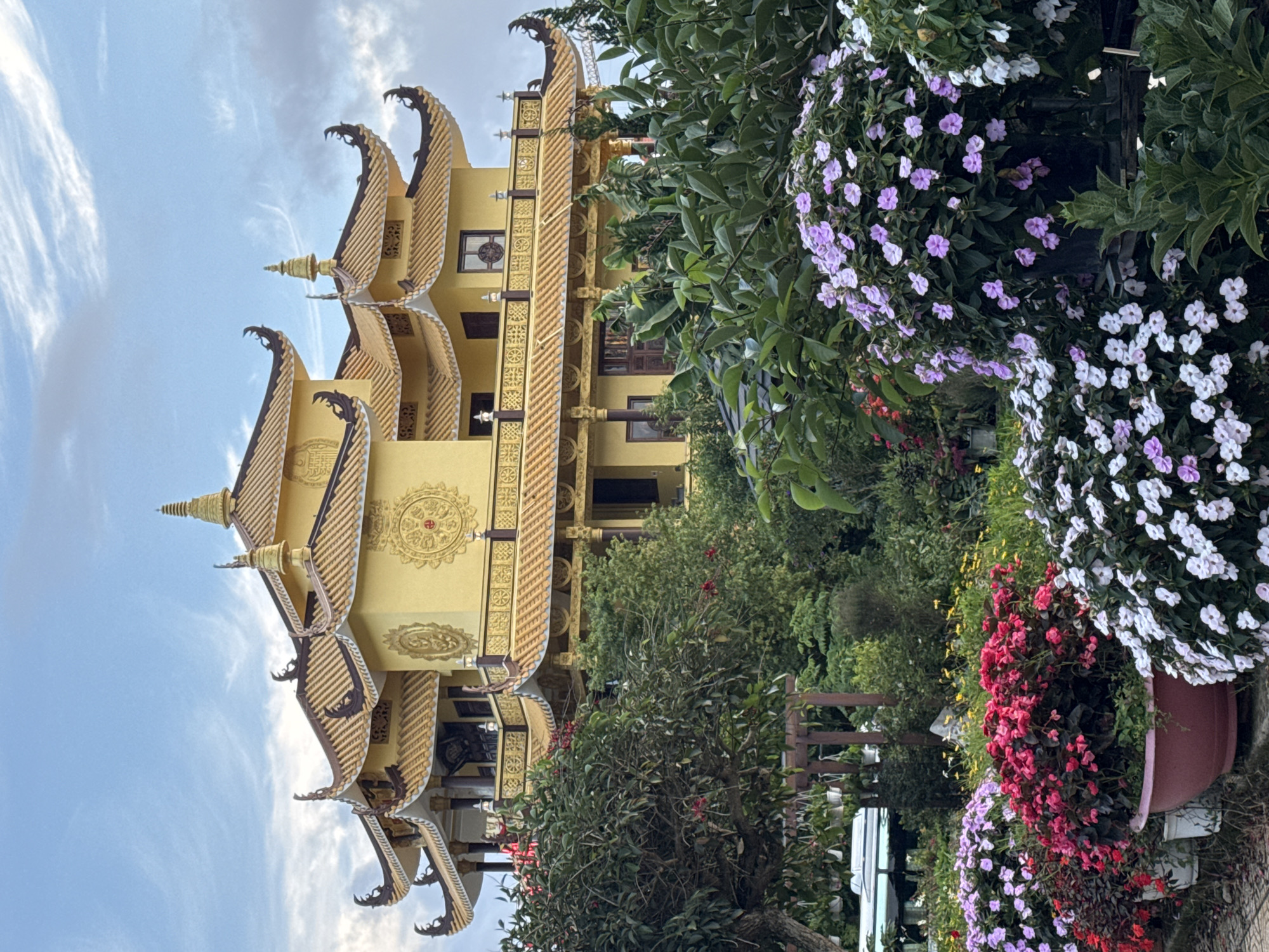 The image shows a Buddhist temple with a yellow, multi-tiered roof, adorned with intricate gold trim and traditional architectural elements. It is situated in the city of Da Lat, Vietnam. In the foreground, a lush garden bursts with vibrant flowers of various colors, including pink, red, white, and purple. Lush green trees and foliage surround the garden, creating a serene and picturesque setting. The sky above is a soft blue with wispy white clouds, suggesting it is daytime. The overall scene conveys a sense of tranquility and natural beauty, characteristic of Da Lat's landscape.