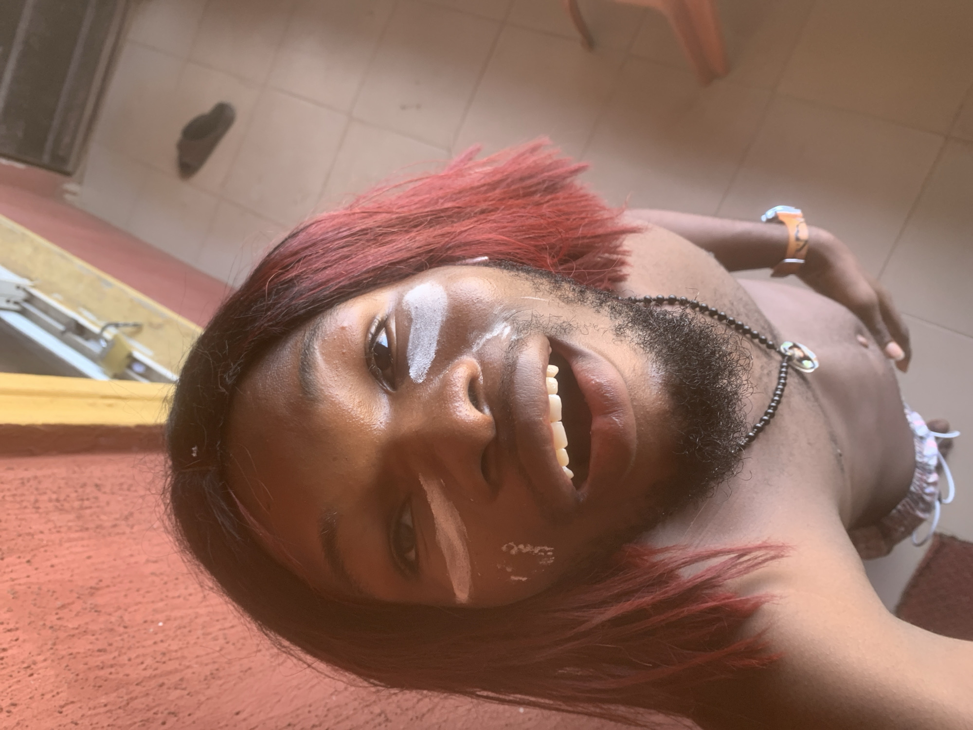 A young person with dark skin, red hair, and a short beard is prominently featured in a close-up, low-angle shot, likely a selfie. They are smiling broadly, revealing white teeth, and looking directly into the camera. White markings, resembling paint or chalk, are visible on their forehead, left cheek, and below their left eye, appearing as lines and dots.
The person is shirtless, exposing their chest and shoulders, and wears a black beaded necklace with a metallic pendant around their neck. An orange-colored wristband or watch is visible on their right wrist.
The background shows a light-colored tiled floor, with what appears to be a dark shoe in the distance. In the foreground, a reddish textured surface is visible, possibly a wall or ground. Natural light suggests it is daytime. The overall mood conveyed is cheerful and expressive, possibly indicating participation in a cultural event or celebration in Nkpor, Nigeria. No discernible text is visible in the image.