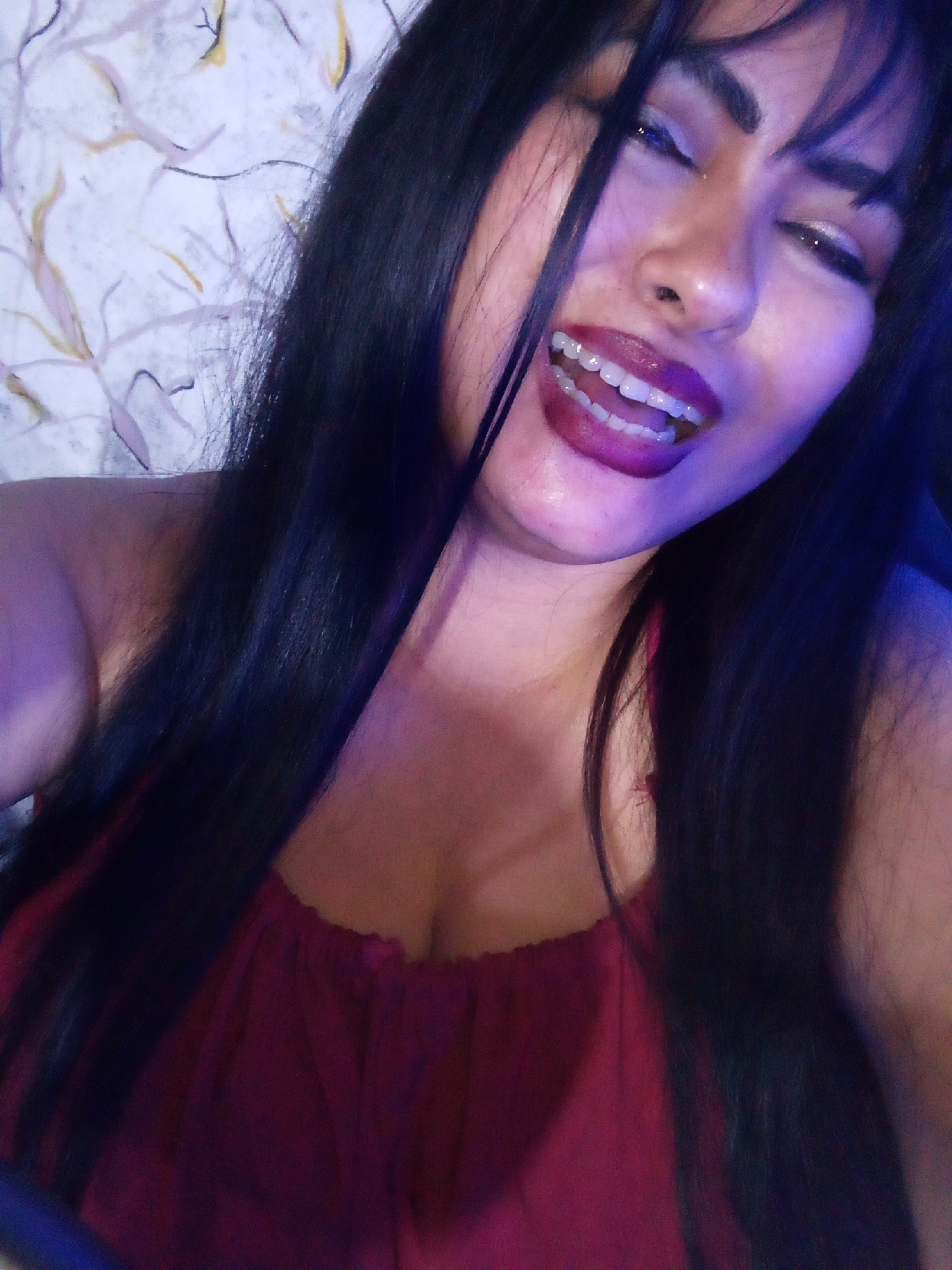 A woman is captured in a close-up, slightly angled selfie. She has long, dark hair that falls over her left shoulder and across the chest. She is wearing a red top with a gathered neckline. The woman is smiling, exposing her teeth, and has dark lipstick on. Her eyes are partially closed, as if she is winking or squinting. The background consists of a patterned wall with light colors and abstract designs. The environment appears to be indoors, and the lighting suggests the presence of a blue light source. The location and city are unknown.