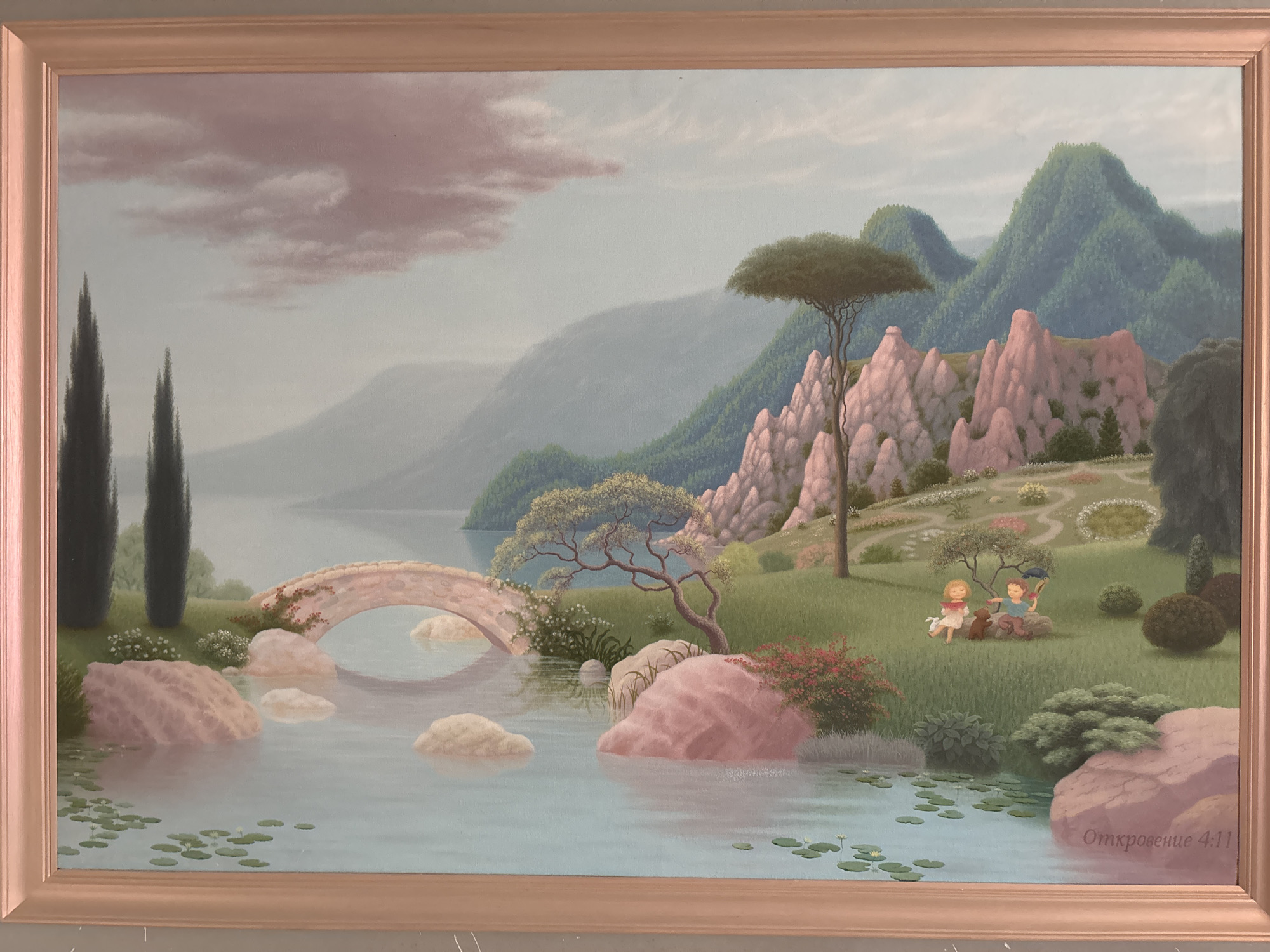 The painting depicts a serene landscape with a body of water in the foreground, featuring lily pads and small yellow flowers. Several large, pink-hued rocks are scattered in and around the water. A stone bridge arches over the water, connecting to a grassy bank. Two cypress trees stand on the left side of the bank, with a cluster of white flowers at their base.
The middle ground of the painting showcases a gentle slope covered in green grass, dotted with small bushes and flowers. A gnarled tree with a sparse canopy stands prominently on this slope. In the background, rolling green hills rise, some with a layered, almost mountainous appearance. The sky above is a pale blue, with soft, pinkish clouds.
On the grassy area in the middle ground, two small children are depicted. One child, in a white dress, stands next to a small brown dog. The other child, in blue, sits on a rock holding a blue umbrella. A pink bush with red flowers is visible in the lower left part of the middle ground.
In the lower right corner of the painting, the inscription "Откровение 4:11" is written in light purple. The painting is framed in a light wooden frame.