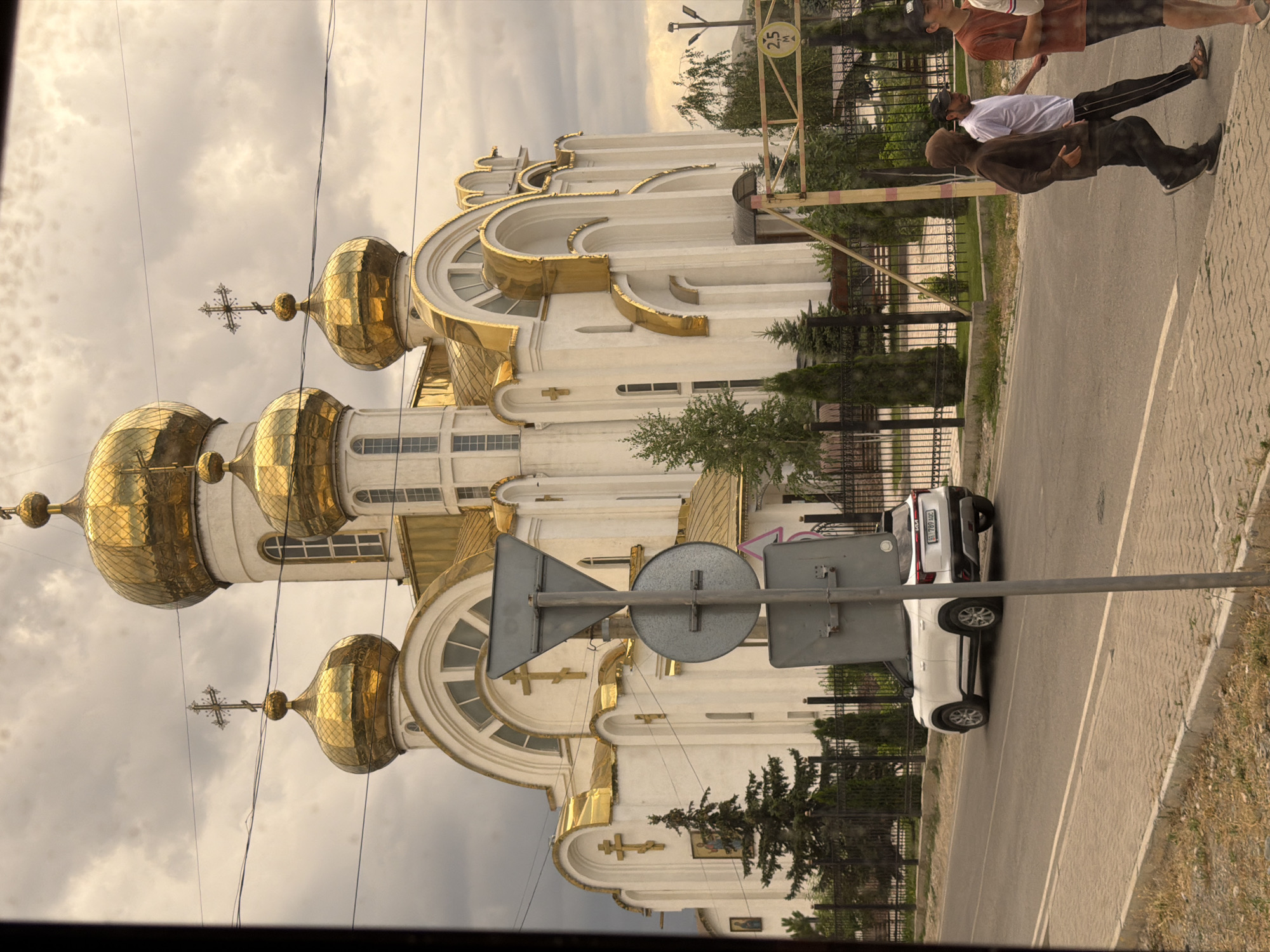 In Unknown, Unknown, a white Kia Seltos is parked on the right side of a paved road, facing left. A large, white building with golden domes and crosses, likely a church, dominates the background. Several people are walking on the sidewalk to the right of the road, some in casual clothing, and others are visible in the distance behind a fence. A traffic signpost with multiple signs stands in the foreground, partially obscuring the view of the car and sidewalk. The sky is cloudy and overcast.