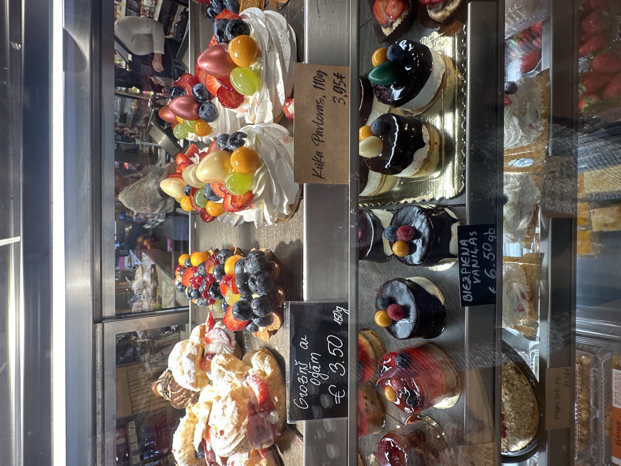 The image shows a display case filled with various desserts. There are several types of cakes and pastries, including pavlovas topped with fruit, tarts filled with berries, and glazed cakes. The prices are displayed on small signs next to each dessert. One sign reads "Kuka Pavlovas, 110g 3,95€," another reads "Groziņš au ogam €3.50 150g," and a third reads "BIEZPIENA VANILAS €6.50 gb." The display case appears to be located in Krustiņi, Latvia.