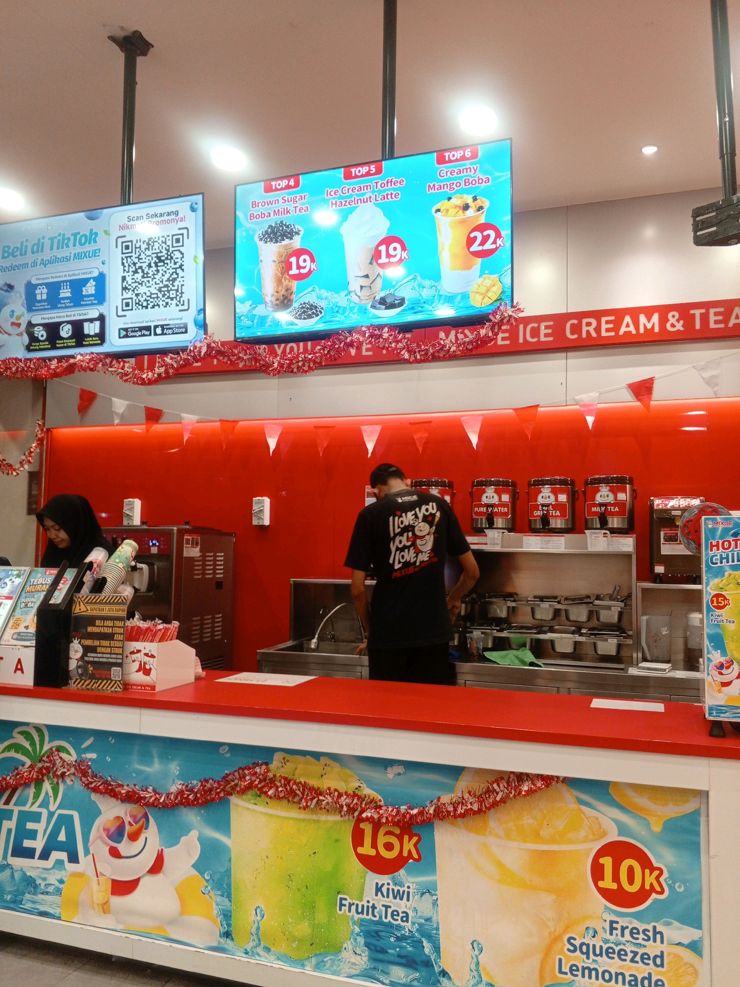 The image shows the interior of a Mixue ice cream and tea shop, likely located in Gabus, Indonesia, as indicated by promotional materials in Indonesian. The scene captures the counter area with two employees. On the left, a female employee wearing a black hijab is visible behind the counter, attending to customers or preparing drinks. On the right, a male employee in a black t-shirt with text "I LOVE YOU YOU LOVE ME" is also working behind the counter.
Above the counter, large digital screens display advertisements for Mixue products. One screen shows "TOP 4 Brown Sugar Boba Milk Tea" for 19k and "TOP 5 Ice Cream Toffee Hazelnut Latte" also for 19k. Another screen advertises "TOP 6 Creamy Mango Boba" for 22k. Further to the right, a sign for "HOT CHILI TEA" priced at 15k is visible.
The counter itself is a bright red, with various beverage dispensers and equipment behind it. There are also decorative garlands in red and white, suggesting a festive or seasonal promotion. In the foreground, a large banner displays graphics of drinks, including "TEA," a snowman-like character, and a cup labeled "16k Kiwi Fruit Tea," and another cup labeled "10k Fresh Squeezed Lemonade." The overall environment appears to be a busy retail space focused on serving cold beverages.