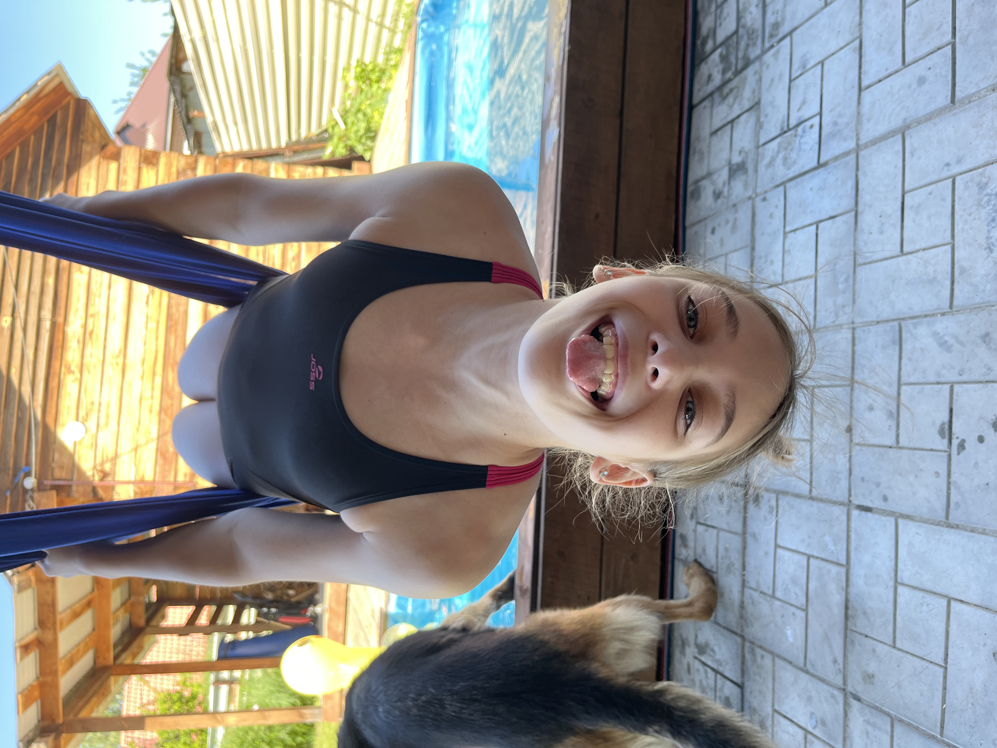 A young Caucasian woman with blonde hair is in the process of training on aerial silks. She is wearing a black and pink swimsuit. Her face is contorted in a playful expression, with her tongue sticking out and her eyes wide open, looking directly at the camera. Her body is positioned in a way that suggests she is suspended in the air, with her hands gripping the blue silks.
The setting appears to be outdoors, possibly a backyard or a training area. Behind her, there is a blue swimming pool, a wooden fence, and some vegetation. The ground is paved with light-colored tiles. A dog, likely a German Shepherd, is partially visible in the foreground, with its back to the camera.
The lighting suggests it is daytime, with bright sunlight casting shadows. The overall mood is casual and energetic, with the woman's expression conveying a sense of fun and playfulness. The scene suggests a warm, sunny day, suitable for outdoor activities. There is some faint text visible on the swimsuit, possibly a brand name. The location context is Novosibirsk, Russia.