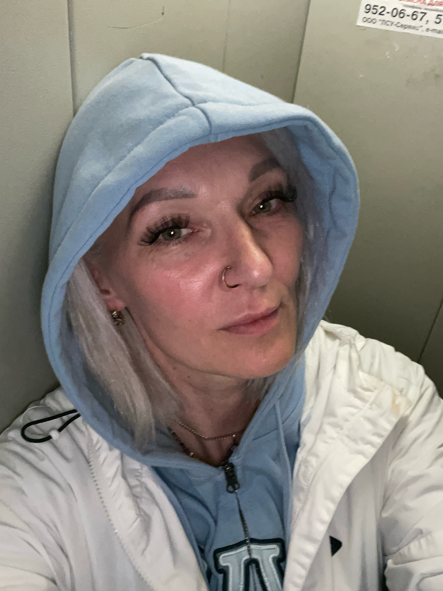 A head and shoulders, close-up, selfie style, portrait of a fair-skinned woman with blonde hair, wearing a light blue hooded sweatshirt and a white jacket. The woman has green eyes, visible eyelashes, and a nose ring. She is looking slightly to her right. The background is a light-colored, textured wall, possibly an elevator or a similar enclosed space. There is a faint sticker or sign on the wall in the upper right corner of the image, with some text in Russian that is partially legible, including a phone number. The lighting is neutral, and the image quality is fair. The photograph was taken indoors.