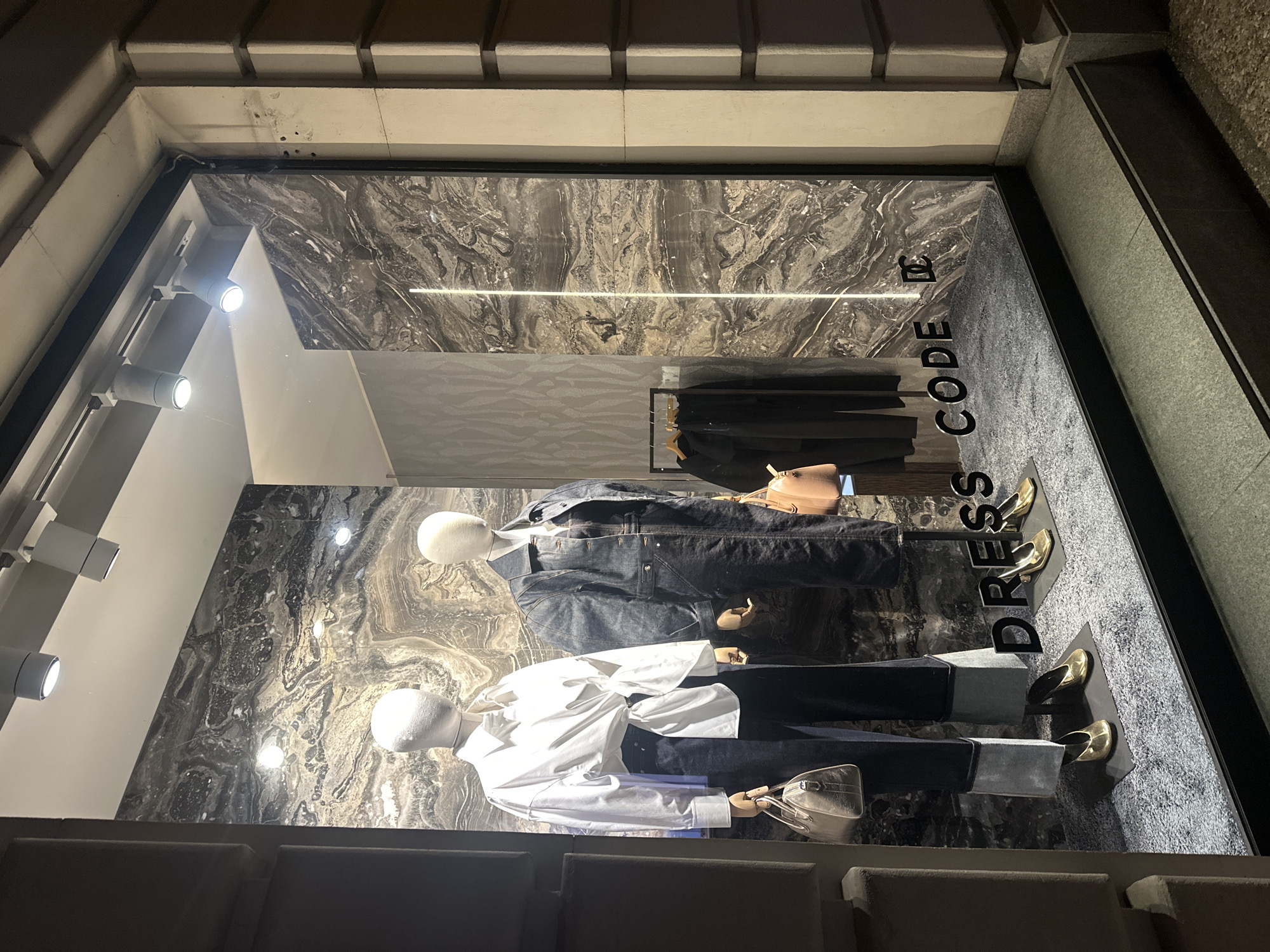 A brightly lit storefront window displays two mannequins dressed in contemporary fashion. The mannequin on the left wears a white oversized shirt with a knot at the waist and dark wide-leg pants, accessorized with a light-colored handbag and gold heels. The mannequin on the right is dressed in a dark denim jumpsuit, also with gold heels. In the background, a dark marble wall features a vertical LED light strip. The floor of the display is covered with a gray, textured rug, and the words "DRESS CODE" are displayed in large black lettering across the front of the display. To the right, a rack of black clothing hangs in the background. The storefront is situated in an urban environment, with a building exterior visible on the right side of the image, featuring light-colored stone architecture.