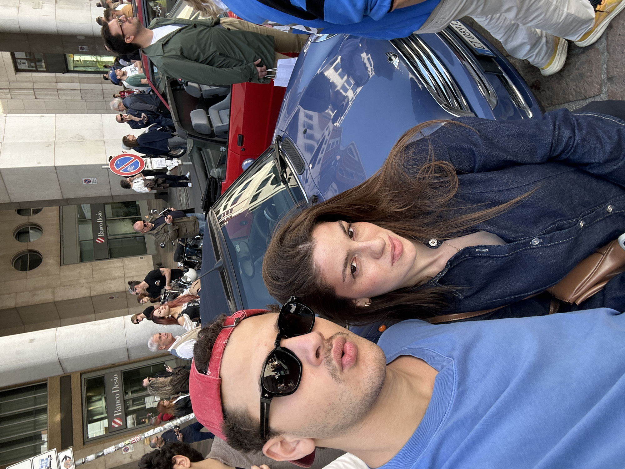 A street-level portrait of two individuals, a man and a woman, possibly a couple, standing beside a blue and red Mini Cooper convertible. The man is in the foreground, wearing a blue t-shirt, a red baseball cap, and black sunglasses. The woman is behind him, wearing a dark denim shirt and a brown leather shoulder bag. Behind them, a crowd of people is gathered, seemingly observing something. A street sign is visible, and a building bearing the sign "Banco Desio" indicates the location is in Milan, Italy.