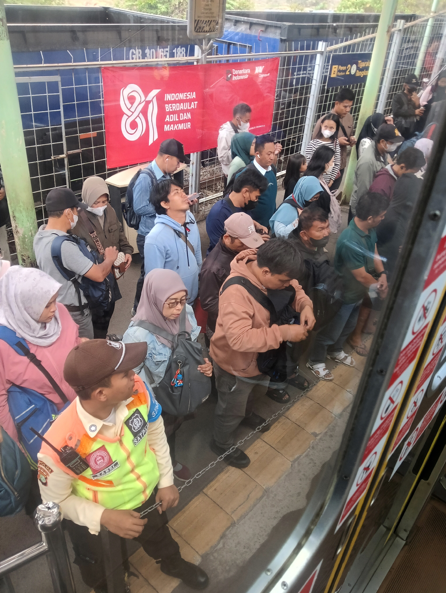 The image captures a crowded scene at a train station in Daru, Indonesia. A diverse group of people, dressed in casual attire and some wearing face masks, are queuing. Some individuals are looking at their phones, while others are engaged in conversation. Several people are carrying backpacks and bags. A security guard, wearing a high-visibility vest over a beige uniform with a walkie-talkie, stands near the front of the crowd, holding onto a chain barrier. Behind the people, a red banner with white text and the number "81" is displayed. The environment appears to be an outdoor platform with a metal fence and a train visible in the background. The overall impression is one of orderly waiting and movement within a public transportation hub.