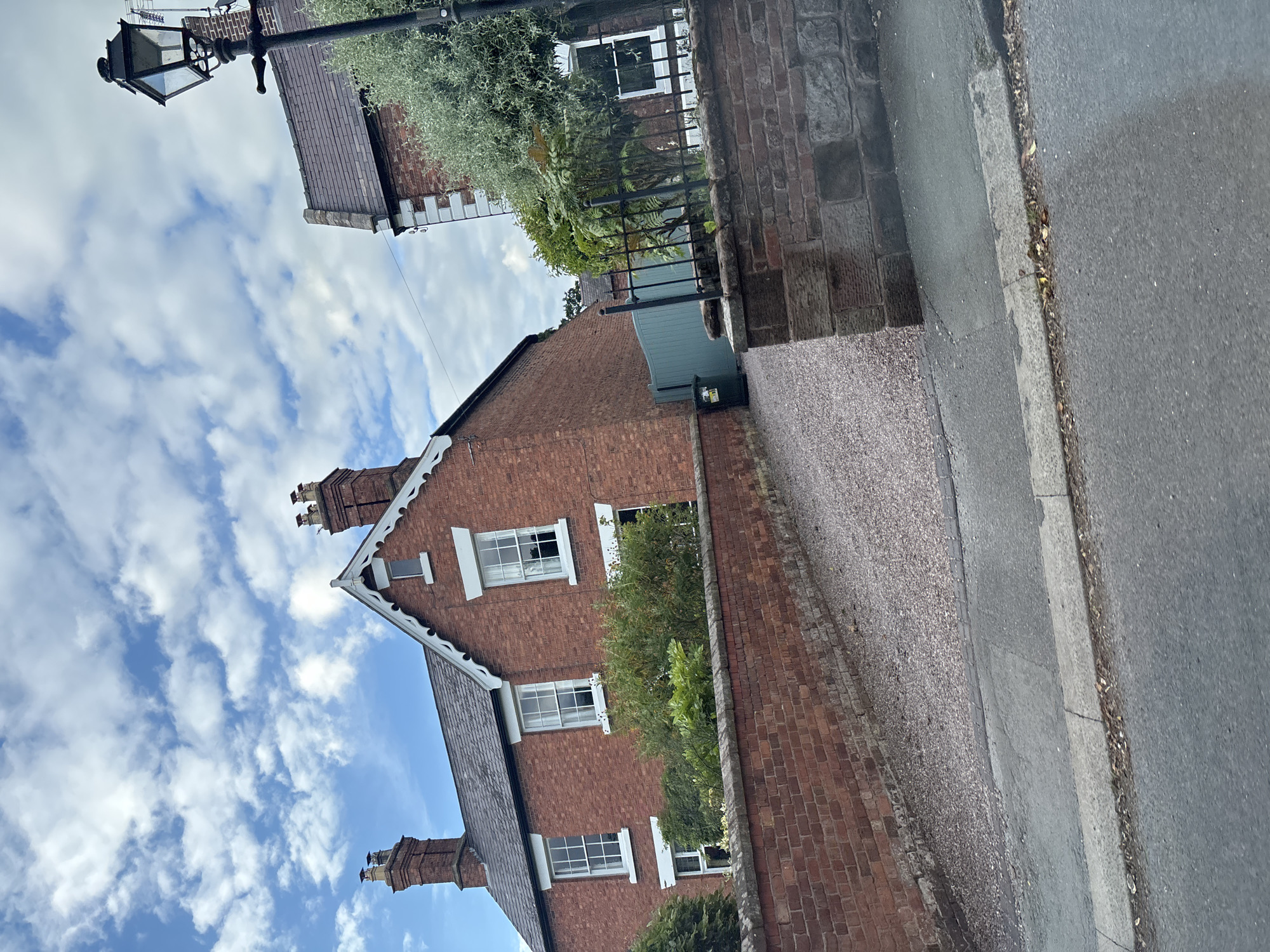 The image shows a red brick house with white-trimmed windows and chimneys. A gravel driveway leads to the house, bordered by a brick wall. There is a lamppost on the right side of the image, and a pale blue gate and a green bin are visible behind the wall. The sky is cloudy. The scene is likely a residential street in Chester, United Kingdom.