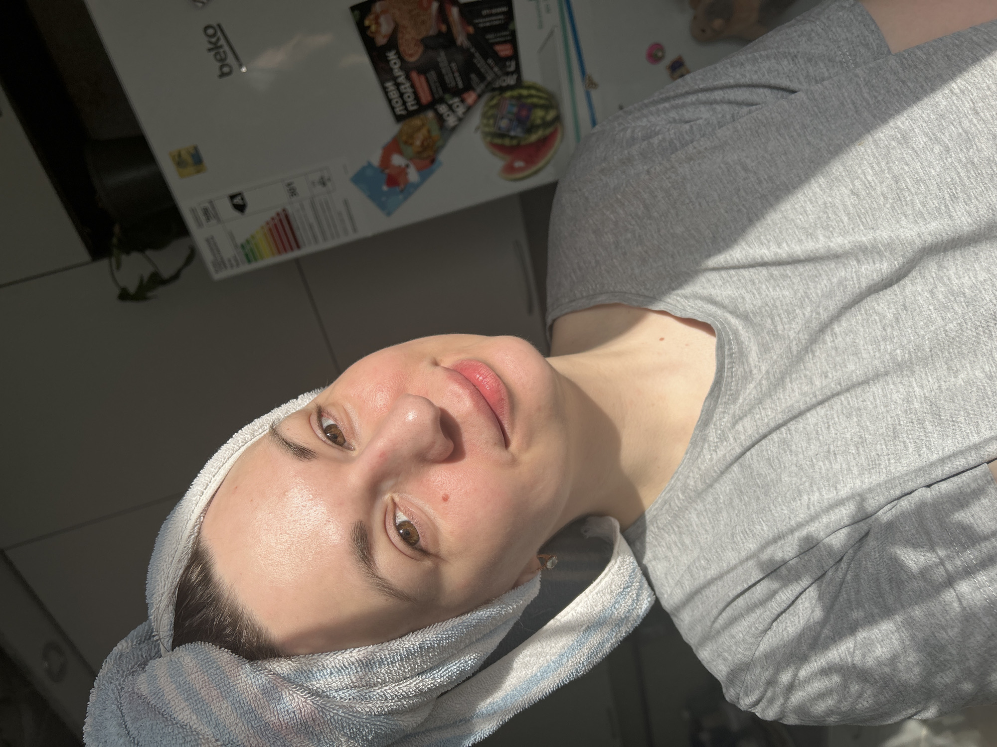A woman with fair skin, hair wrapped in a white and blue striped towel, is wearing a gray t-shirt. The photo is taken indoors with artificial lighting. In the background is a white refrigerator with stickers on it. Based on the refrigerator brand "beko," the image may not be in Фоки, Russia.
