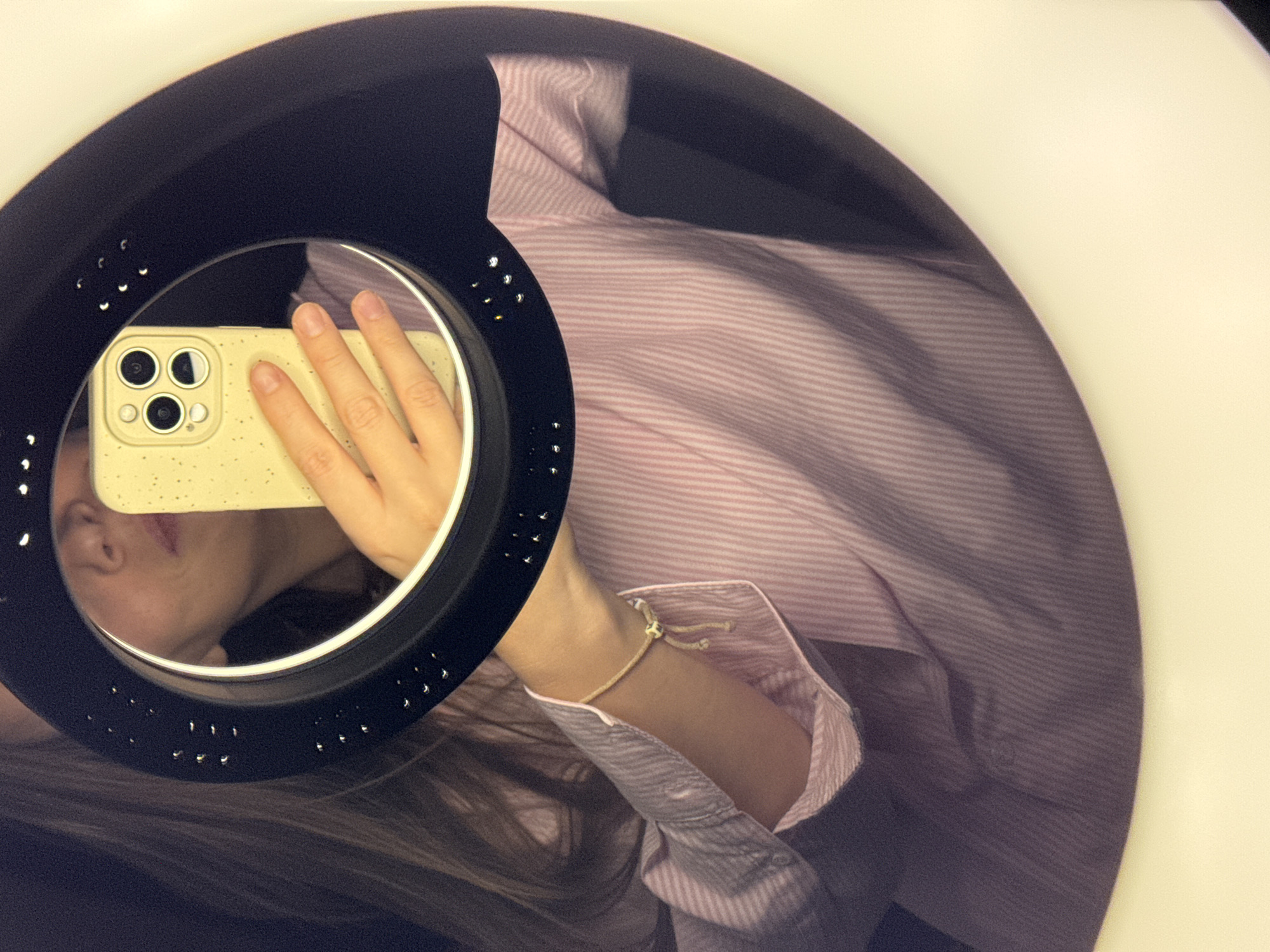 The image is a close-up, slightly tilted selfie taken in a mirror illuminated by a ring light. The mirror reflects a person holding a smartphone with a beige case. The person is wearing a pink and white striped collared shirt with the sleeve casually rolled up to reveal a thin, beige cord bracelet. The ring light is black with evenly spaced circular holes around its inner circumference. The reflection shows part of the person's face, including the nose and lips. Their hair is visible, cascading down. The environment appears to be indoors. There is no evidence in the image to support a location in the city of Budva, Montenegro.