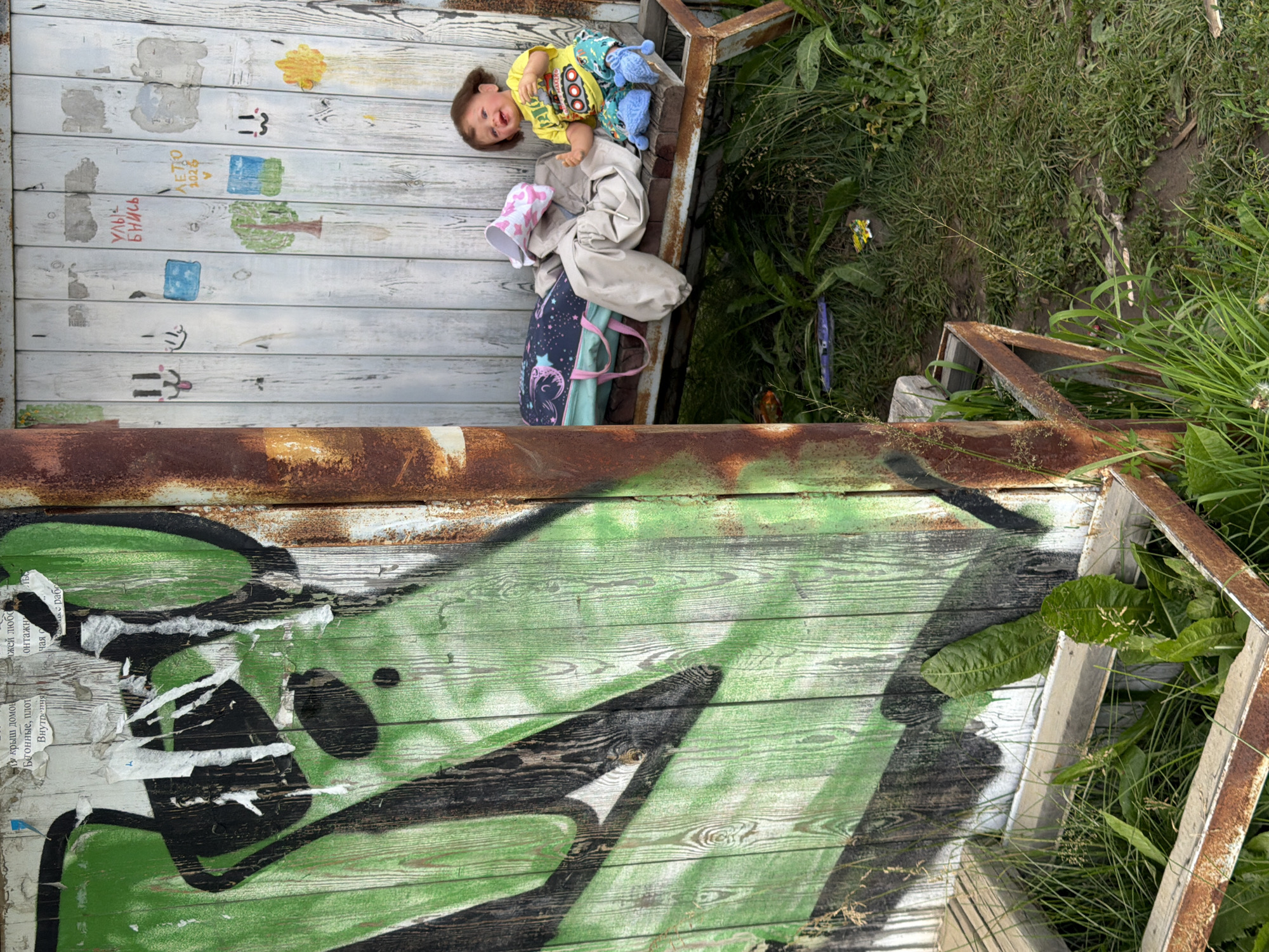 A young child, appearing to be a doll, sits on a weathered wooden bench against a white wooden wall covered in peeling posters and children's drawings. The doll wears a yellow t-shirt and patterned pants, with blue slippers. A backpack and pile of clothing are on the bench beside the doll. To the left, a rusted metal pole stands in front of a wooden surface covered in vibrant green graffiti. The ground is covered in grass and weeds, with some discarded items visible. The scene is located in Zheleznodorozhnyi, Russia.