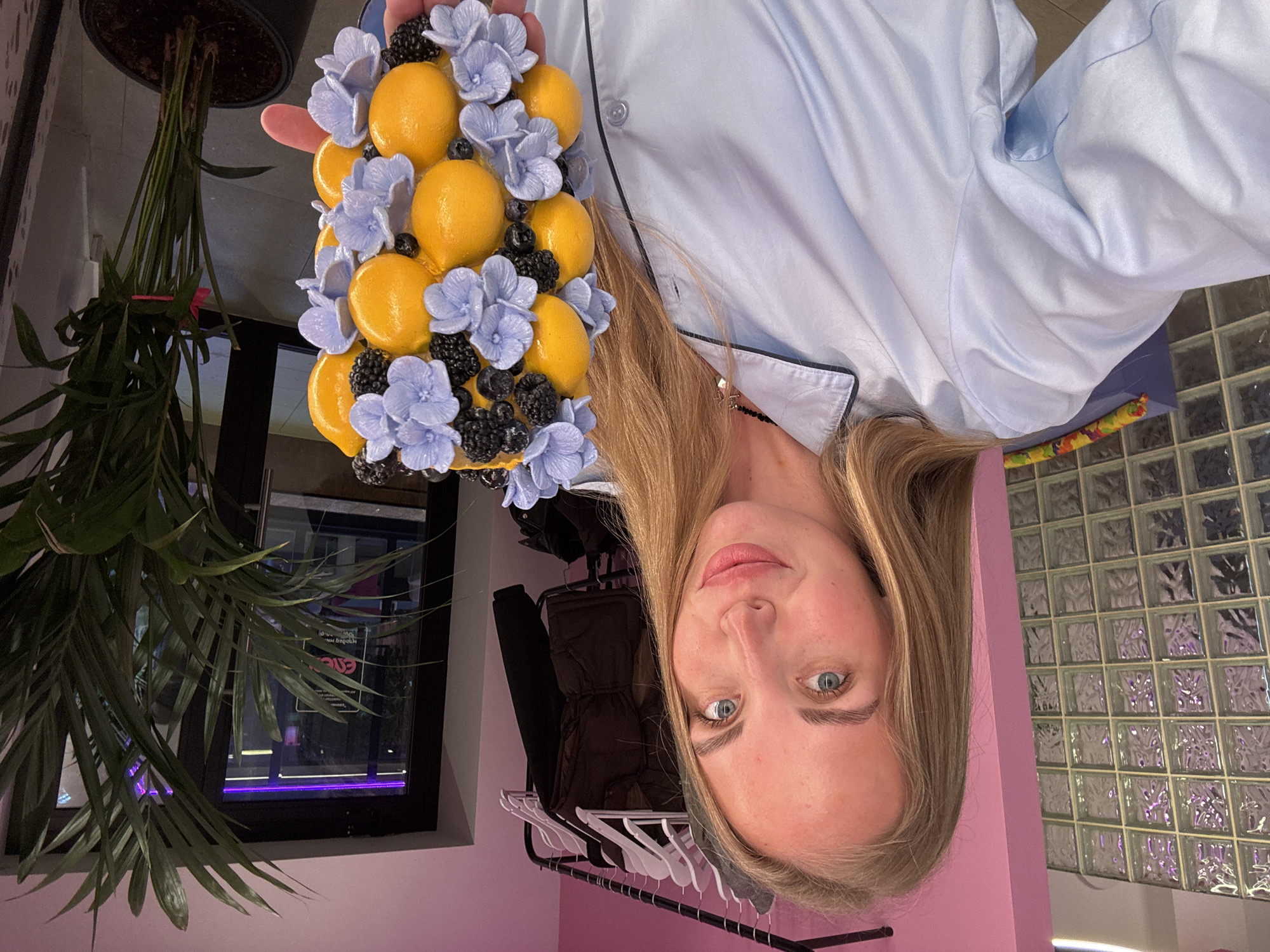 A fair-skinned woman with long blonde hair is holding a decorative sphere composed of lemons, blueberries, blackberries, and light blue flowers in her left hand. She is wearing a light blue long-sleeved pajama top with black piping. The background includes a potted palm plant near a window, a clothes rack with clothing, a pink wall, and a wall of textured glass blocks. The location appears to be indoors, possibly a home or a retail space. There is no explicit information about the location being in Sochi, Russia within the frame.