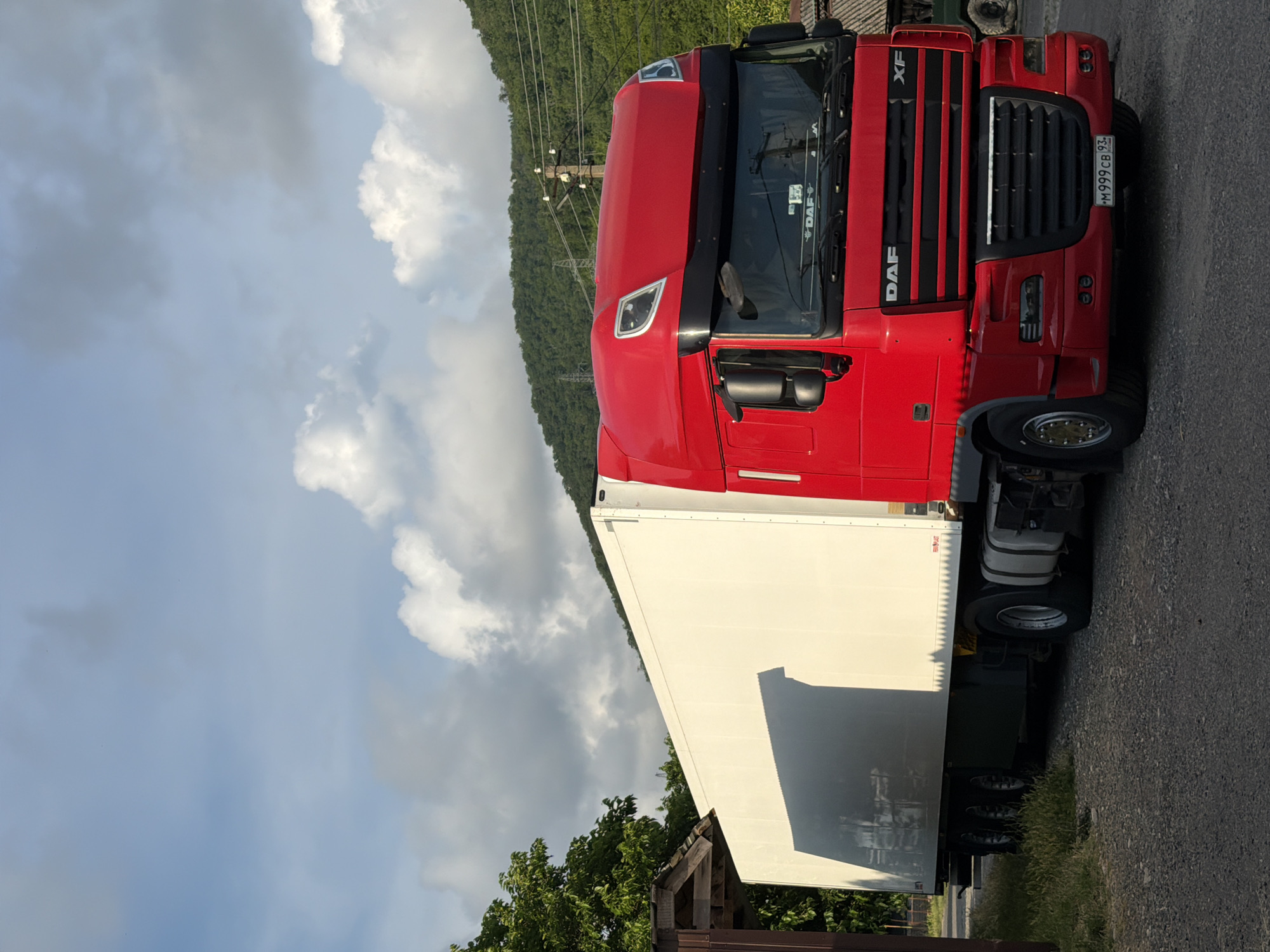 This is a landscape-oriented photograph of a bright red DAF semi-trailer truck parked on a gravel road in Shepsi, Russia. The truck has a white trailer attached and is oriented vertically in the frame, as if the camera is tilted. The foreground shows the truck's tires and the road surface, which is a mix of gravel and asphalt. The middle ground features the truck itself, with its distinctive red cab and large white trailer. The truck's front grille and headlights are visible, as is the "DAF" logo on the side of the cab. A license plate with Cyrillic lettering, "M 999CB 93", is attached to the front of the truck.
In the background, a lush green, tree-covered hillside rises steeply. Power lines and poles are visible against the hillside, suggesting the proximity of infrastructure. The sky is a mixture of blue sky and dramatic, fluffy white clouds, indicating a bright day with some cloud cover. The lighting suggests it is daytime, likely late afternoon given the shadows. The overall scene conveys a sense of a parked vehicle in a rural or semi-rural setting, possibly on a route through hilly terrain. There are no people visible in the image.