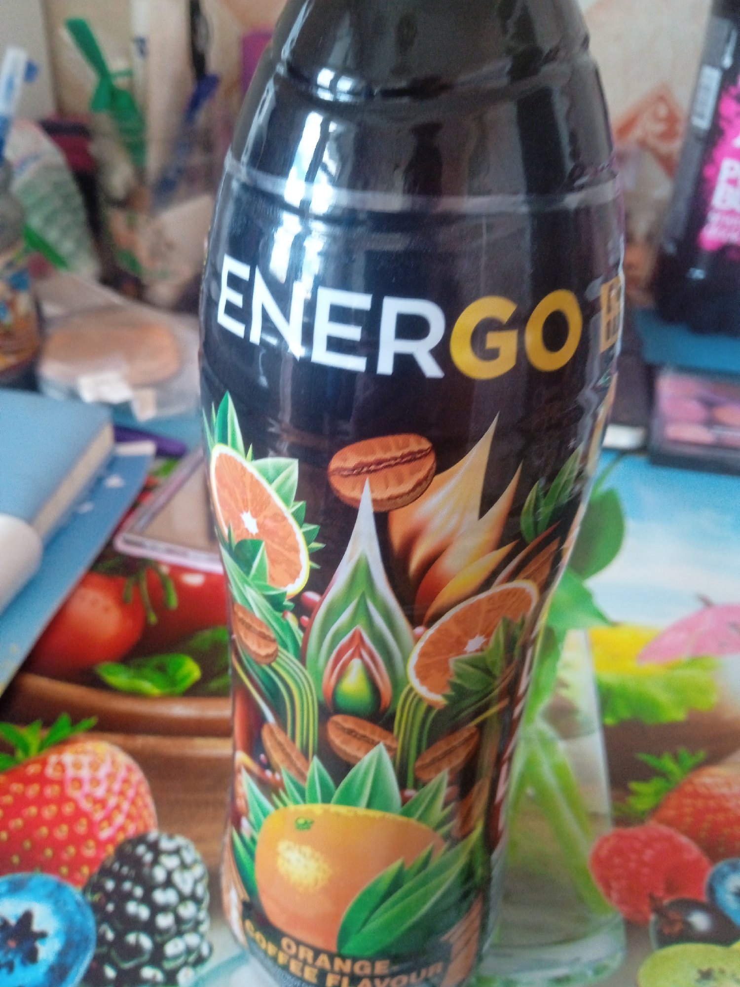 The image shows a close-up of a bottle of "ENERGO" brand beverage, likely an energy drink. The bottle has a black label with white and yellow lettering. The label features illustrations of fruits, coffee beans, and green leaves. Specifically, there are depictions of orange slices, strawberries, blueberries, and blackberries. The text "ORANGE COFFEE FLAVOUR" is visible at the bottom of the label.
The bottle is standing on a surface that appears to be a table or counter, with other objects in the blurred background. These objects include what looks like other bottles of drinks, some packaging, and possibly some books or papers. The overall lighting suggests an indoor environment. There is no indication of the specific location within the city of Chernivtsi, Ukraine. The image is a still photograph and does not depict any action or interaction.