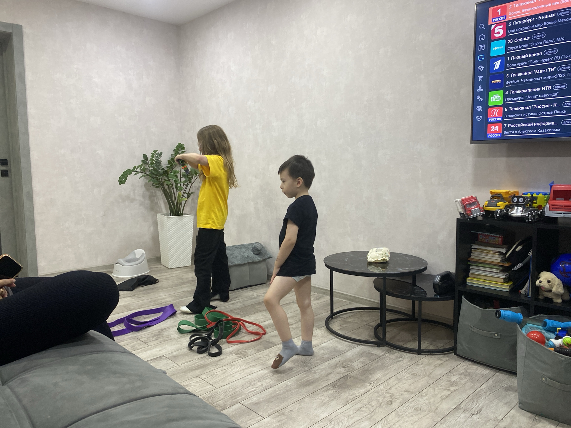 Two children, a girl and a boy, are in a room with light-colored walls and a wood-patterned floor. The girl, wearing a yellow t-shirt and black pants, stands by a potted plant. The boy, in a black t-shirt and grey socks with a bear design, stands facing away from the camera. Several resistance bands in purple, green, and red are on the floor near the children. A small white potty is also on the floor. To the right, a black round coffee table with a white object on it sits next to a black shelving unit filled with books and toys. A large TV screen displays a television guide. A person is partially visible on a gray couch on the left side of the image, holding a mobile phone. The scene suggests a domestic setting.