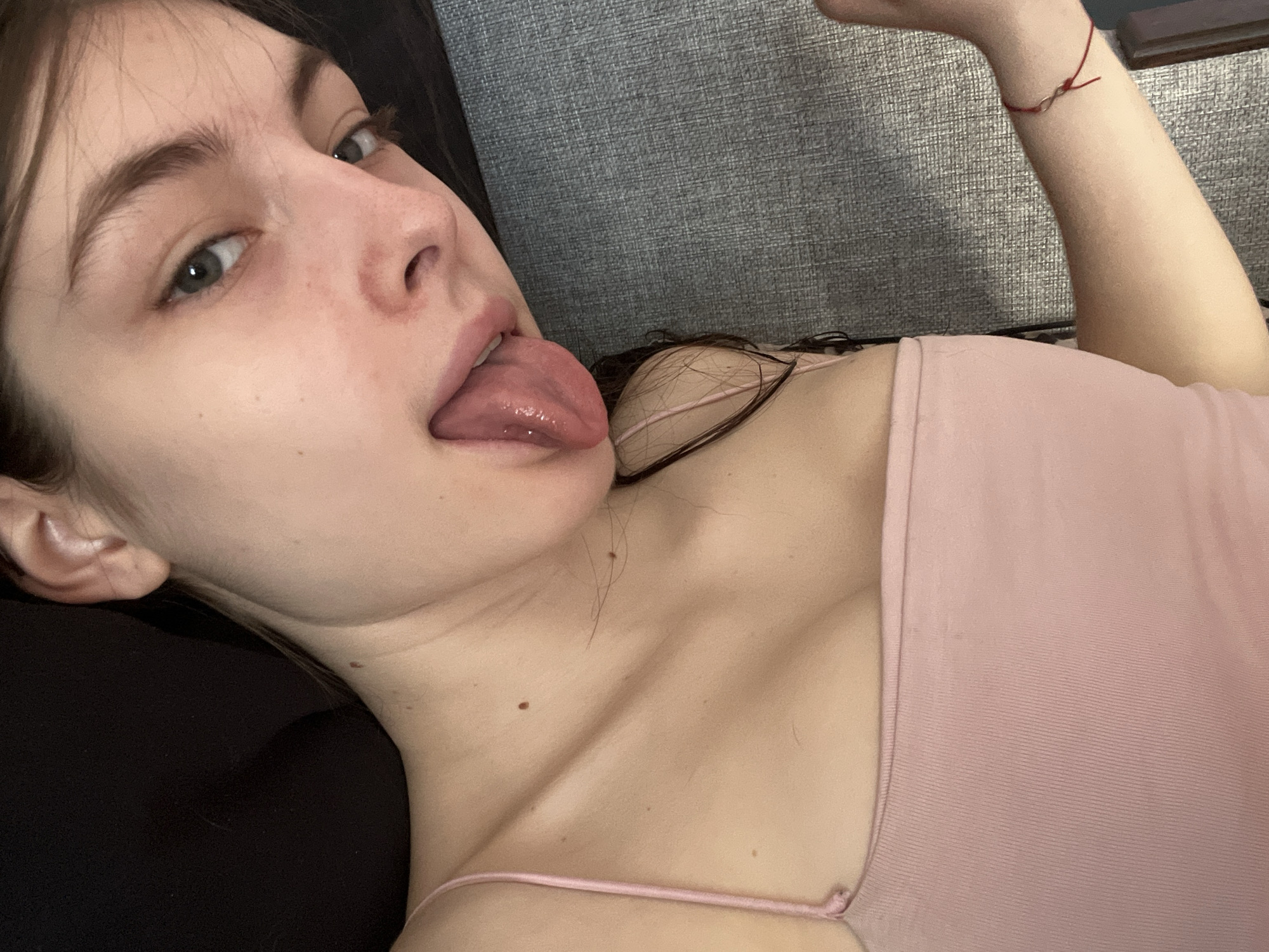 A young woman lies on a surface with gray fabric, likely a couch or bed. She has fair skin, dark hair, and green eyes. Her tongue is sticking out. She is wearing a light pink tank top with thin straps. A red bracelet is visible on her wrist. The background is blurred, but it appears she is indoors. There is no evidence of Saint Petersburg, Russia.