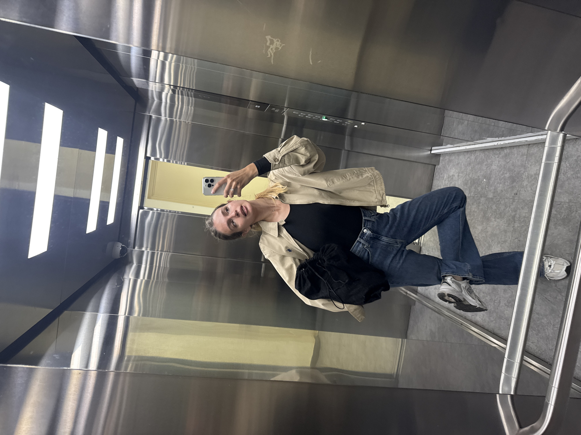 The image shows a young woman taking a selfie in an elevator. She is wearing a black top, blue jeans, and a beige jacket. She is holding a smartphone in her right hand. The elevator appears to be made of stainless steel. There is a handrail on the right side of the elevator. The location is Unknown, Unknown.
