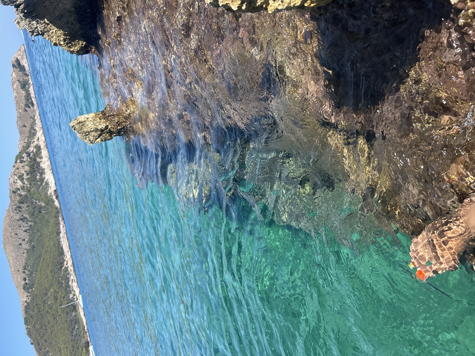 The image shows a close-up view of clear, turquoise water lapping against a rocky shoreline. A person's bare foot, partially submerged in the water, is visible in the lower left corner. The foot appears to be resting on the submerged rocks. The water is very clear, allowing the rocky bottom to be seen. In the distance, a hillside covered in green vegetation slopes down to the water. A white sailboat is anchored in the blue sea. The sky above is a clear, pale blue. The overall impression is a tranquil and scenic coastal environment. The location is identified as Cala Lliteres, Spain.