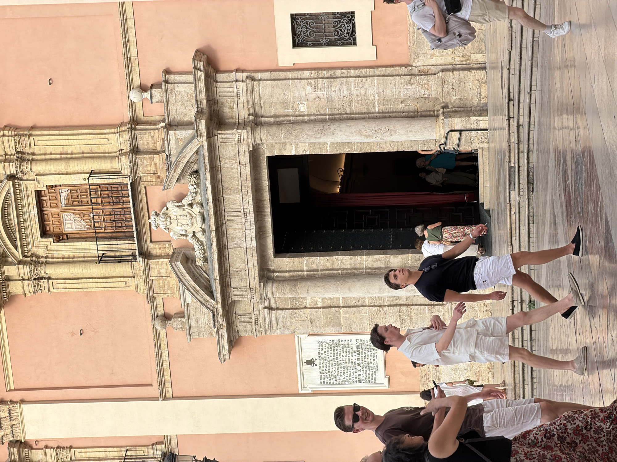 In an outdoor, urban setting, a group of people are walking near a large, ornate building with a pink facade. In the foreground, a woman with dark curly hair wearing a dark sleeveless top and a floral skirt walks from left to right. She is followed by a man wearing sunglasses, a dark t-shirt, and white shorts. To the right of the man in sunglasses, a younger man in a white linen shirt and white shorts walks briskly. Further to the right, another young man in a black t-shirt and white shorts walks with a determined stride, carrying sunglasses on his shirt. Behind them, other individuals are visible, including someone carrying a large backpack. The building features a grand entrance with large stone columns and intricate carvings. A plaque is mounted on the facade to the left of the entrance. The ground is paved with large stone tiles that reflect the light. Several steps lead up to the building's entrance. The overall scene suggests a warm, daytime environment.