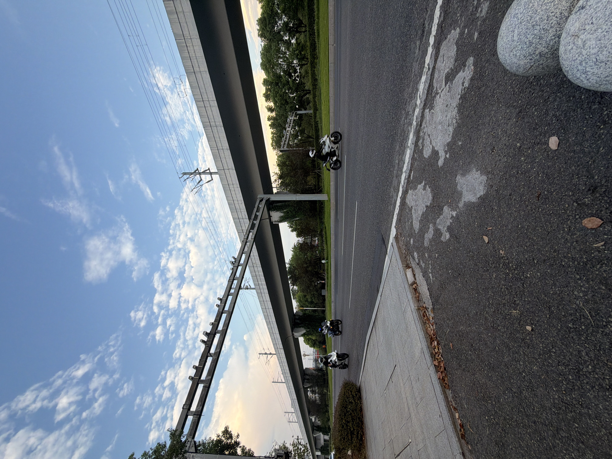 In the city of Jiangjin, China, a scene unfolds beneath an elevated structure, likely a railway or highway overpass. The sky is a blend of blues and soft whites, with wispy clouds suggesting either early morning or late afternoon. The overpass, a substantial concrete construction, casts a shadow over the asphalt road where two motorcycles are in motion. The first motorcycle, predominantly white, is ridden by an individual wearing a white helmet and dark clothing. It is positioned on the left side of the road, heading away from the viewer. Slightly further ahead and to the right, a second motorcycle, also white, is being ridden by a person in a blue helmet and dark attire, moving in the same direction.
The road surface is asphalt, with a visible white lane divider. To the left of the road is a paved sidewalk, distinguished by grey paving stones. Along the edge of the sidewalk, a scattering of dry leaves indicates the presence of nearby trees, which are visible as a dense line of green foliage beyond the road. The overall environment suggests an urban or semi-urban setting, characterized by modern infrastructure and natural elements. The lighting indicates a time of day with diffused sunlight, creating a calm and tranquil atmosphere.