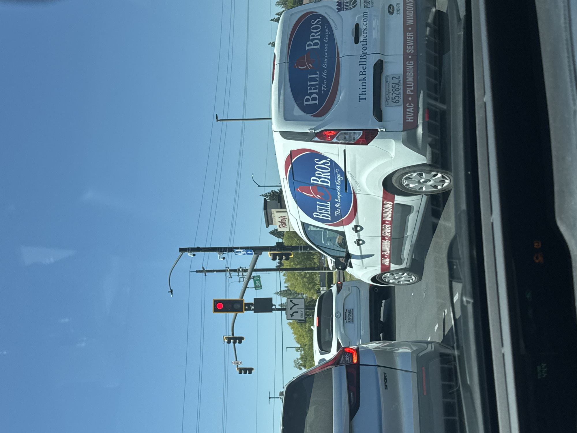 The image shows a daytime, outdoor view from a car looking forward at traffic. The vehicles are stopped at an intersection with a red traffic light. A white Ford van with "Bell Bros." branding is in the right lane. The van's side and rear display logos and text indicating services like HVAC, plumbing, sewer, and windows, along with the website "ThinkBellBrothers.com." The van has a California license plate that reads "65285L2." In the center lane, a white Mazda SUV with license plate "8ZF5X84" is stopped. To the left of the Mazda, a silver SUV is partially visible. Street signs indicate "Lichen Dr" and directional arrows. The environment appears to be a suburban area with trees and businesses visible in the background. The sky is clear blue. This scene is located in Citrus Heights, California, United States of America, likely at the intersection of Sunrise Boulevard and Lichen Drive, given the presence of a Raley's store and other commercial businesses typical of the area.
