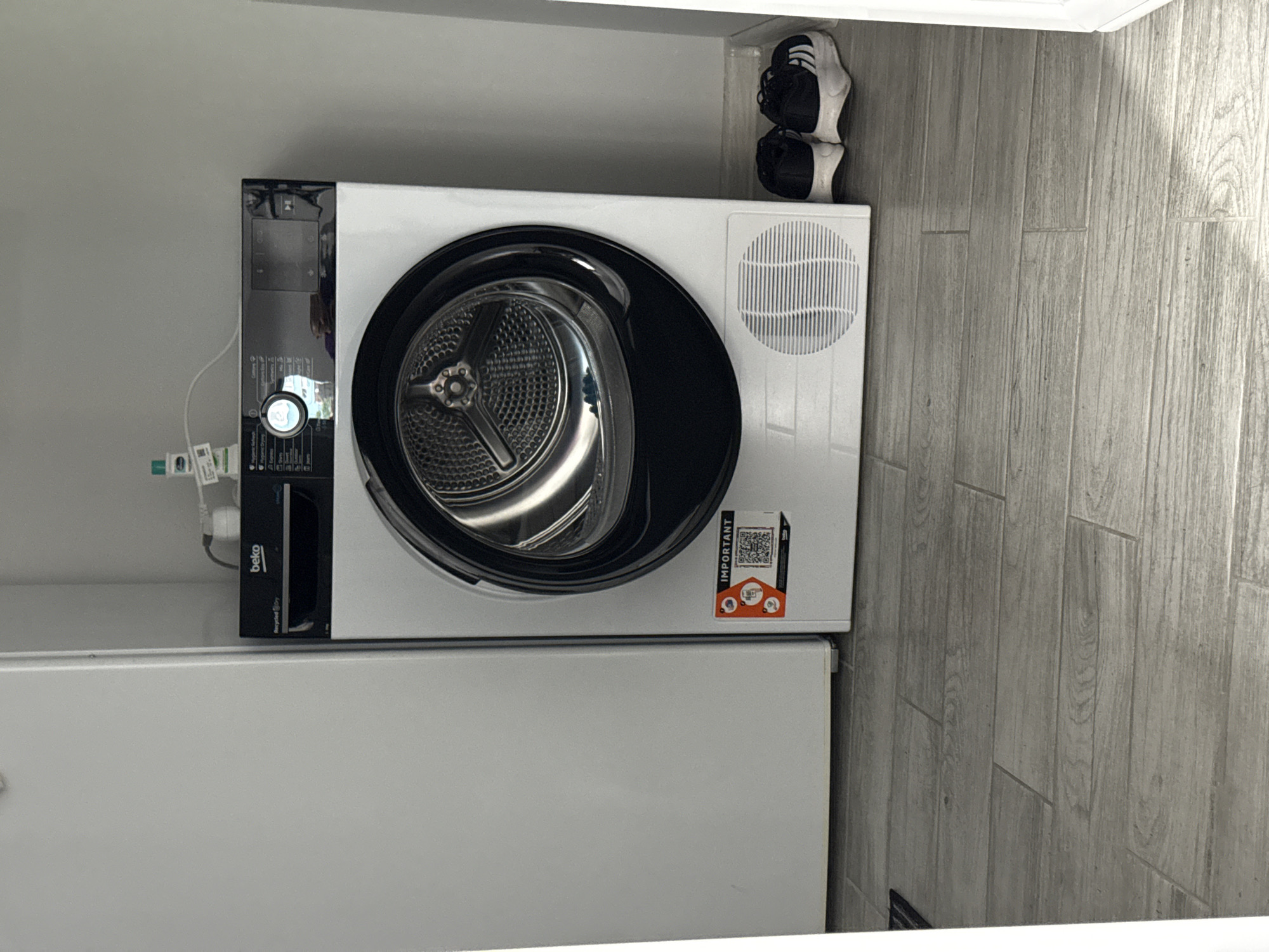 An indoor scene in Stara Zagora, Bulgaria, depicts a white Beko-branded tumble dryer positioned against a light gray wall. To the left, a portion of a tall, white, rectangular appliance, likely a refrigerator, is visible. The dryer features a black control panel at the top with a central illuminated dial displaying various drying programs such as "Hygiene Refresh," "Cotton," "Express," "Synthetics," "Wool," "Jeans," and "Outdoor." The front of the dryer has a large circular glass door revealing a metallic drum interior. A circular ventilation grille is present at the bottom front of the appliance. A rectangular sticker labeled "IMPORTANT" with a QR code and the Beko logo is affixed to the lower right side of the dryer. Above the dryer, a small white bottle with a green label, identified as Cetaphil, rests on the appliance's surface, connected to a power outlet via a white cord. Two pairs of black and white athletic shoes are placed on the tiled floor to the right of the dryer. The floor consists of light-colored, wood-patterned rectangular tiles laid in a staggered pattern. No individuals are present in the image.