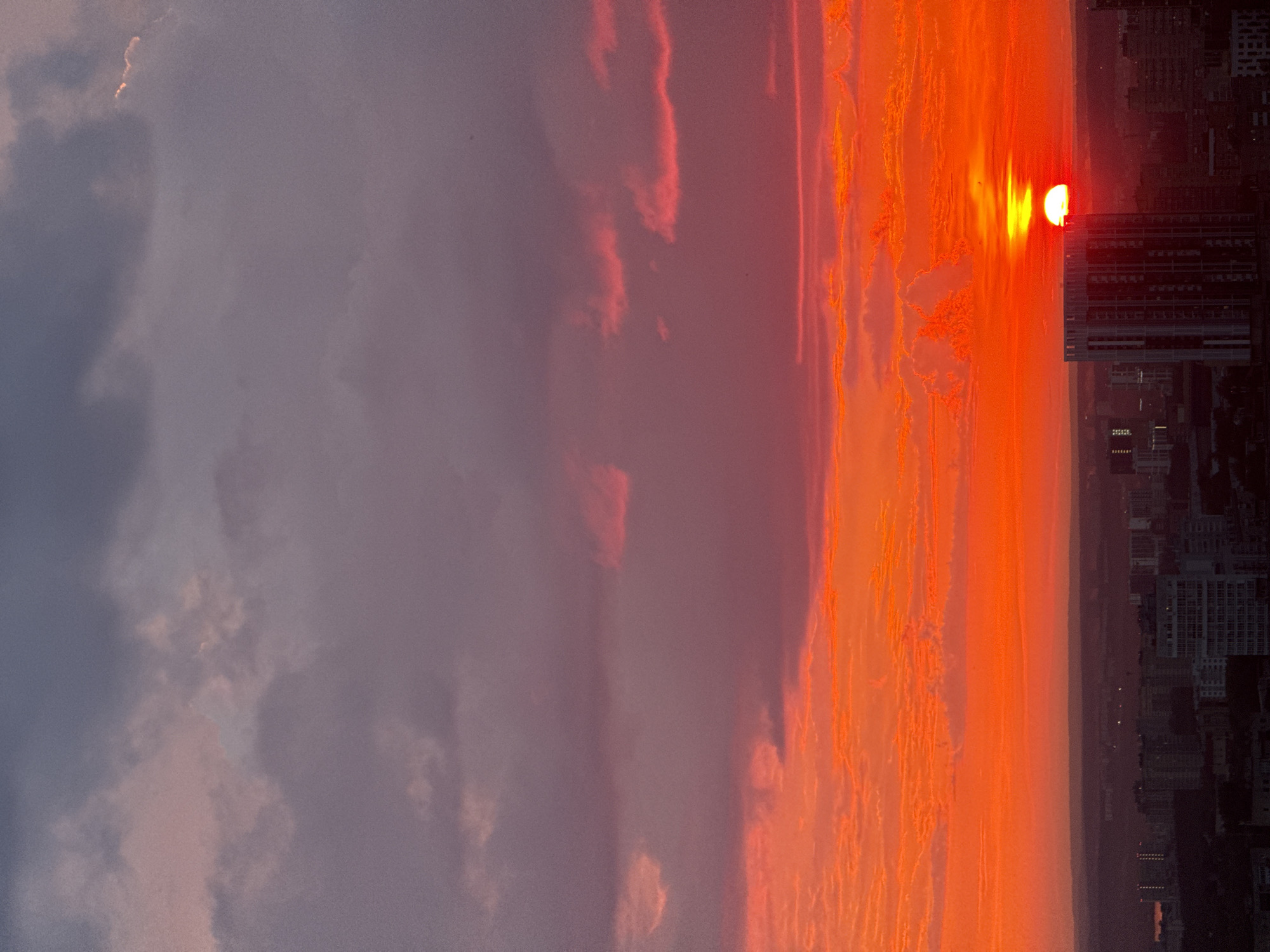 The image depicts a dramatic sunset over a city, likely Savenki, Russia, given the provided context. The sky is ablaze with vibrant hues of orange and red, transitioning to a soft pink and then a deep purple-grey at the top. Wispy clouds are illuminated by the setting sun, creating streaks of light against the darker sky. The sun itself is a brilliant orb, partially obscured by the horizon line, casting a warm glow across the scene.
In the foreground, a silhouette of a city skyline is visible. Numerous buildings of varying heights are discernible, with one taller, modern-looking structure standing out. The cityscape is cast in shadow due to the low angle of the sun. The environment appears to be an urban setting during twilight. There are no visible people or specific actions taking place in the image. The overall impression is one of a natural phenomenon occurring over a populated area.