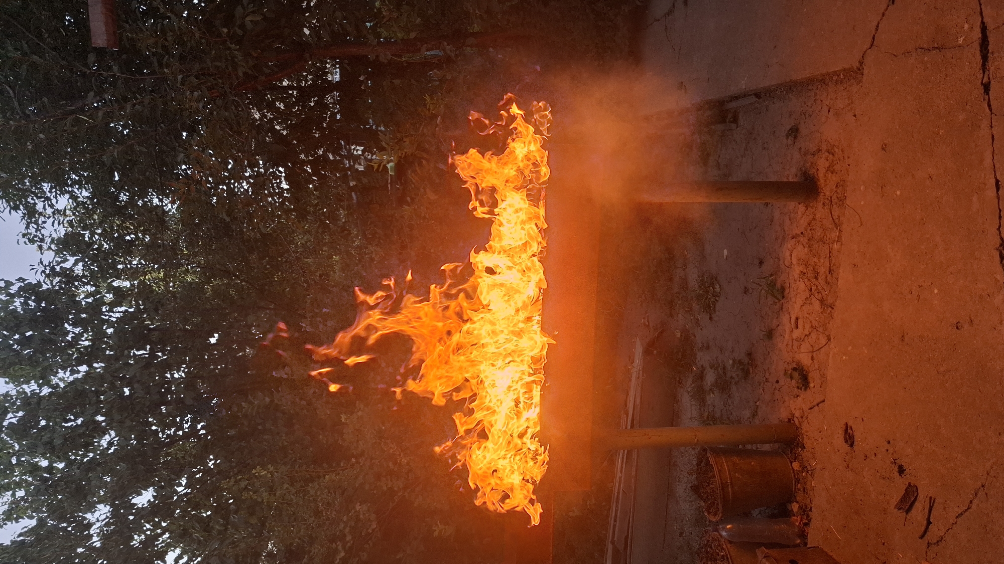 In an outdoor setting, likely a backyard in Шұбарсу, Kazakhstan, a metal grill or fire pit is ablaze with a vibrant orange and yellow fire. The flames are intense and reach upwards, illuminating the surrounding area with a warm glow. Behind the fire, dense green foliage from trees creates a natural backdrop, with the sky visible as a pale, overcast hue. In the foreground, the ground is a mixture of dirt and cracked concrete, with some scattered debris and two buckets visible. One bucket appears to be a metal pail filled with dark material, possibly soil or organic matter, and next to it sits a plastic bottle. The scene captures a moment of active fire, suggesting an outdoor gathering or activity.