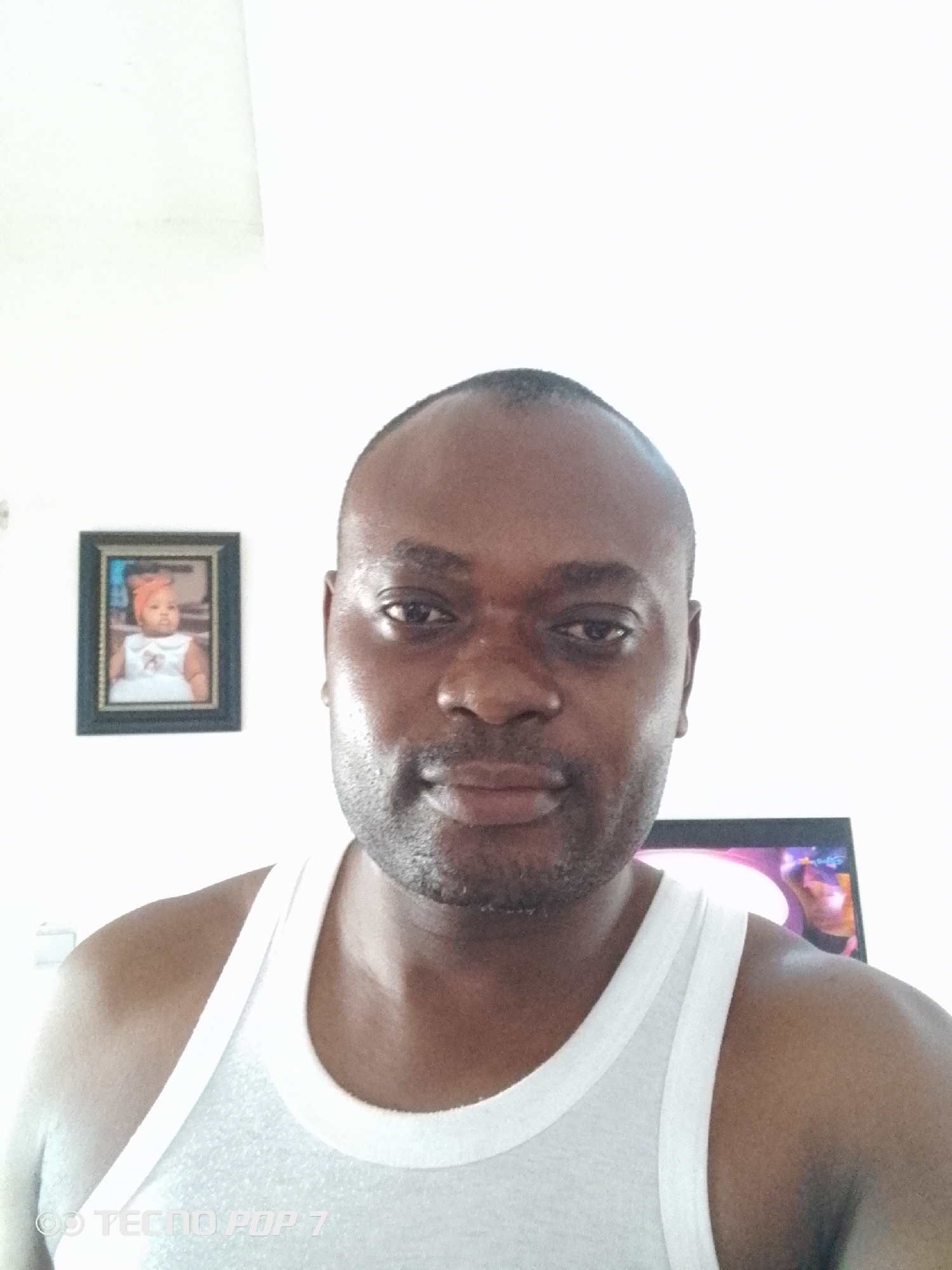 A medium shot depicts a Black man with a closely shaved head, dressed in a white tank top. He is facing the camera with a neutral expression. Behind him, on the white wall, hangs a framed photograph of a baby. Below the photograph and partially obscured, is a television. The location is likely a residence in Abuja, Nigeria. A "TECNO POP 7" watermark is visible in the bottom left corner.