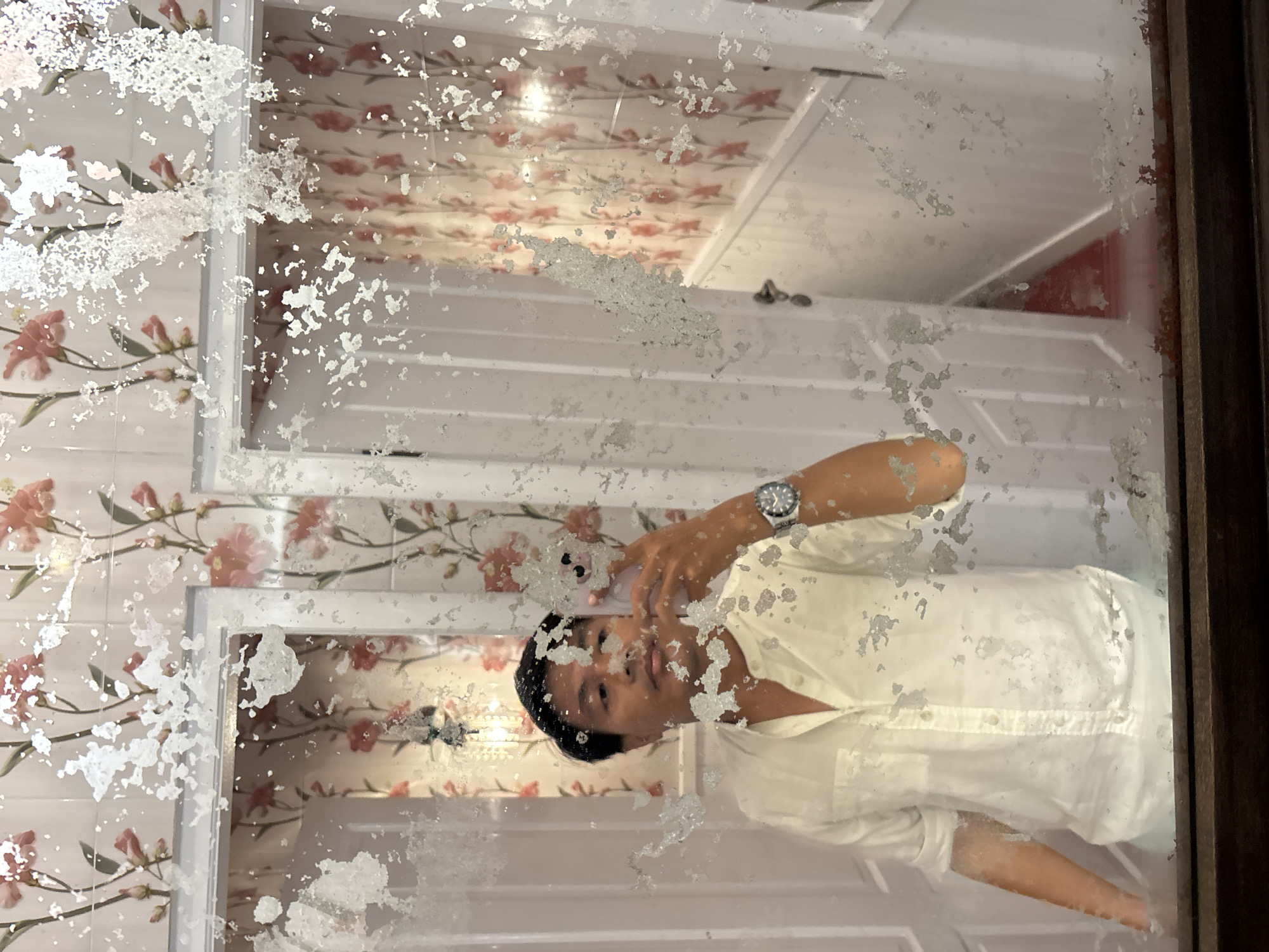 A person takes a selfie in a dimly lit room with floral wallpaper. The mirror is old and has large sections where the silvering has peeled away, obscuring parts of the reflection. The person is wearing a white button-down shirt and a watch. In the reflection, there appear to be two doorways and a red rug on the floor. The room is likely a bathroom or dressing area.