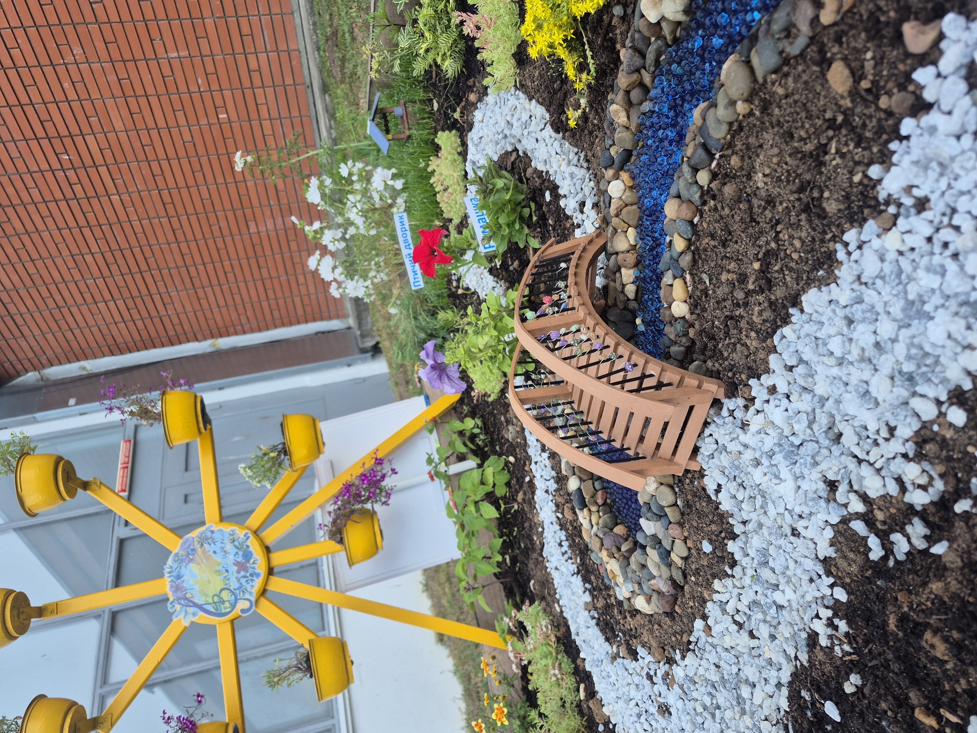 The image depicts a whimsical garden display, possibly a fairy garden, set against a brick wall. The garden features a miniature wooden bridge spanning a decorative "river" made of blue glass beads and pebbles. Alongside the "river" are pathways of white gravel and dark soil, dotted with small plants and flowers, including a vibrant red bloom and purple petunias.
A prominent yellow Ferris wheel-like structure, adorned with small planters, stands to the left of the garden, adding a playful element. Some of the planters contain trailing purple flowers and greenery. In the background, a section of a brick wall is visible, along with some green foliage.
Several small signs with Cyrillic text are placed within the garden. One sign near the red flower reads "Птичий дворик," which translates to "Birdyard." Another sign near a green plant appears to say "Римма."
The overall scene suggests a bright, perhaps sunny, day, given the clear lighting and the vibrant colors of the garden elements. The meticulous arrangement of the stones, beads, and plants, along with the miniature bridge and Ferris wheel, creates an imaginative and charming atmosphere. The location context of Chaikovsky, Russia, provides a regional setting for this outdoor decorative display.
