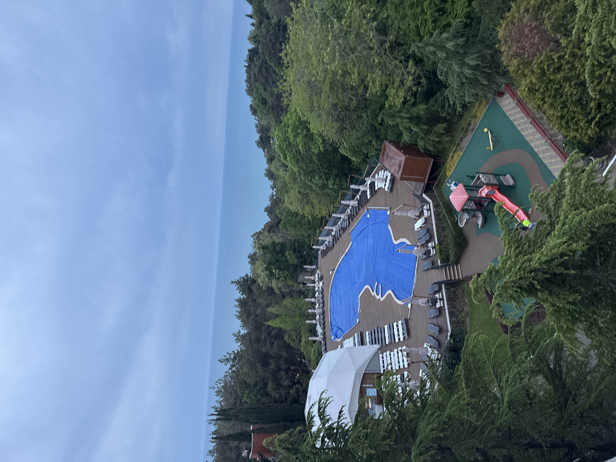 The image depicts a high-angle view of a resort in Sosnovy, Russia. A covered blue swimming pool is surrounded by lounge chairs on a wooden deck. Adjacent to the pool area is a children's playground featuring slides and other play equipment. Lush green trees surround the resort. In the background, a hazy blue body of water meets the horizon under a pale blue sky. A white geodesic dome structure is also visible near the pool.